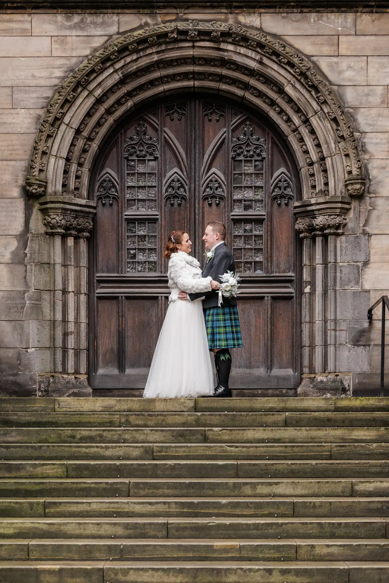 A bride in a white gown and a groom in a green tartan kilt embrace on stone steps before a large arched wooden door with ornate carvings and gothic windows, set in a stone building.