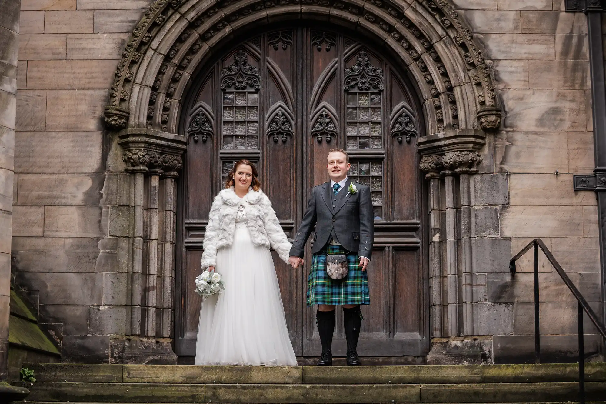 A bride in a white dress and fur jacket stands holding hands with a groom in a green tartan kilt and jacket. They stand on stone steps in front of an ornate, arched wooden church door with detailed carvings.