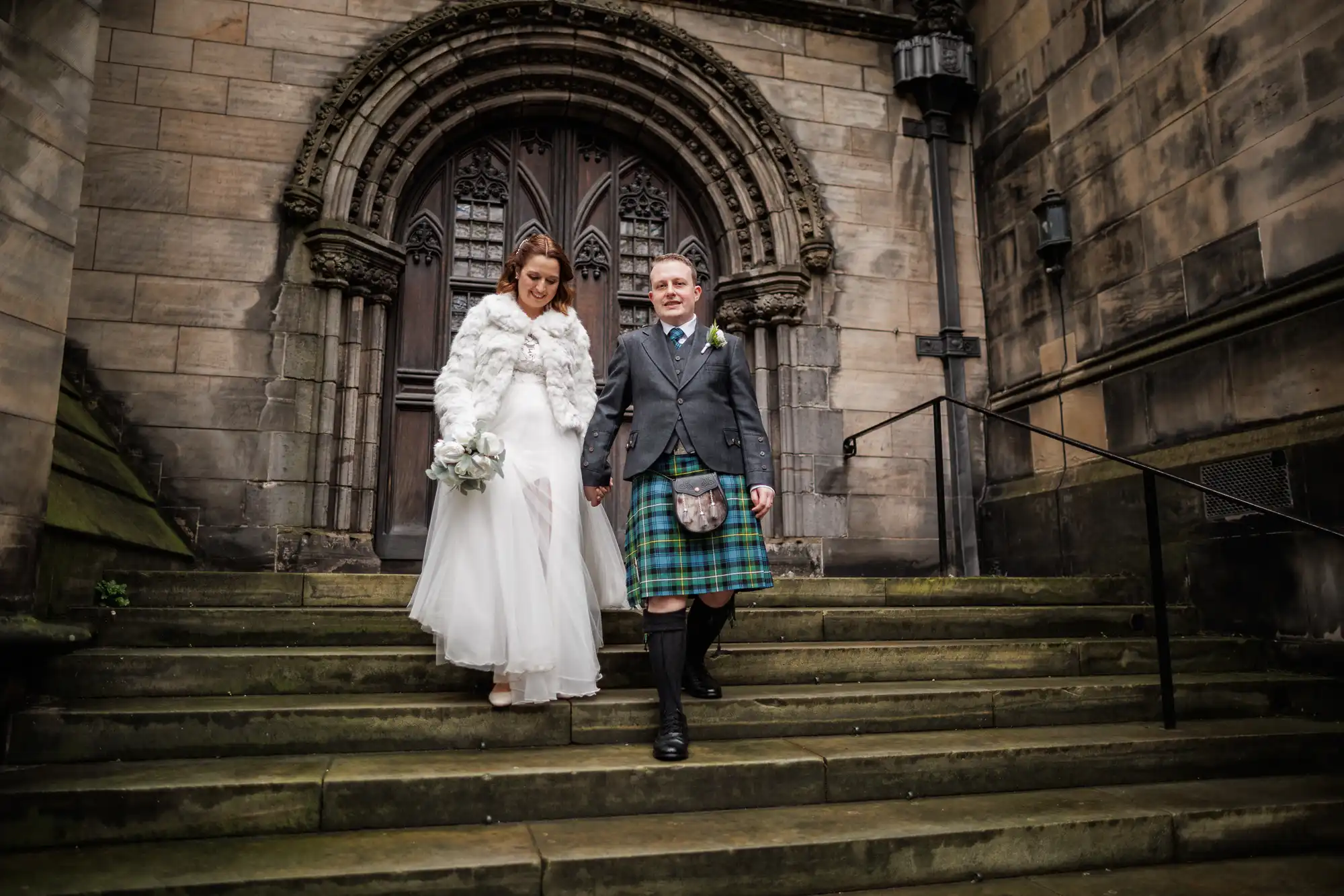 A bride in a white dress and fur shawl walks down stone steps beside a groom in a grey jacket, green tartan kilt, and sporran. They hold hands and smile, with a bouquet in her hand. Behind them is a large, ornate wooden door set in a stone archway.
