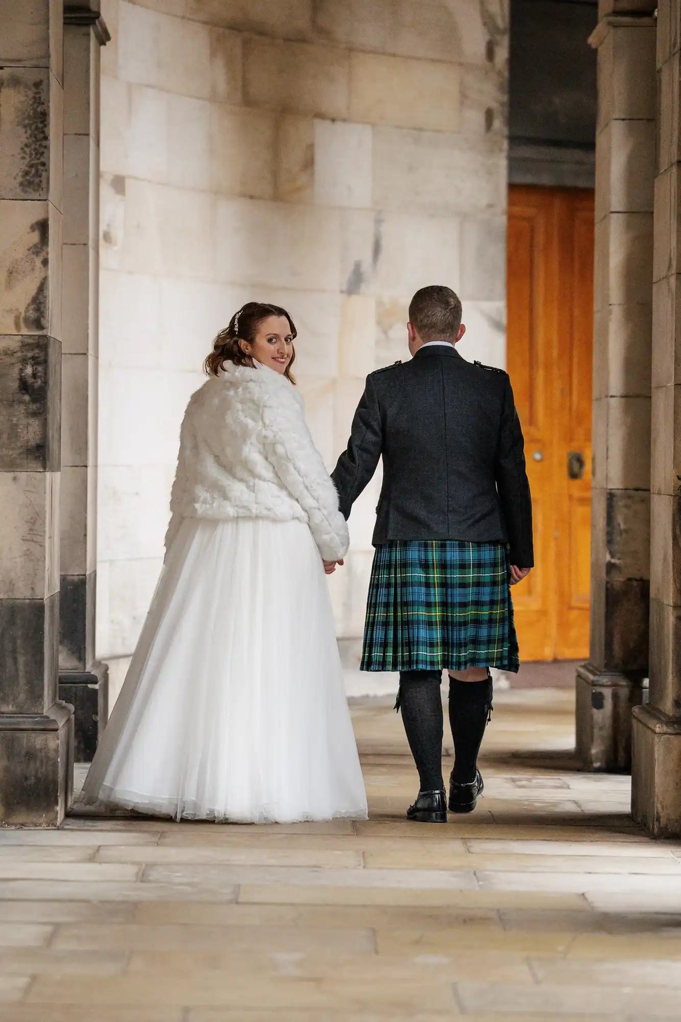 A bride in a white dress and faux fur jacket holds hands with a groom in a dark jacket and green tartan kilt. They walk away down a stone corridor, looking back and smiling, with a bright wooden door in the background.
