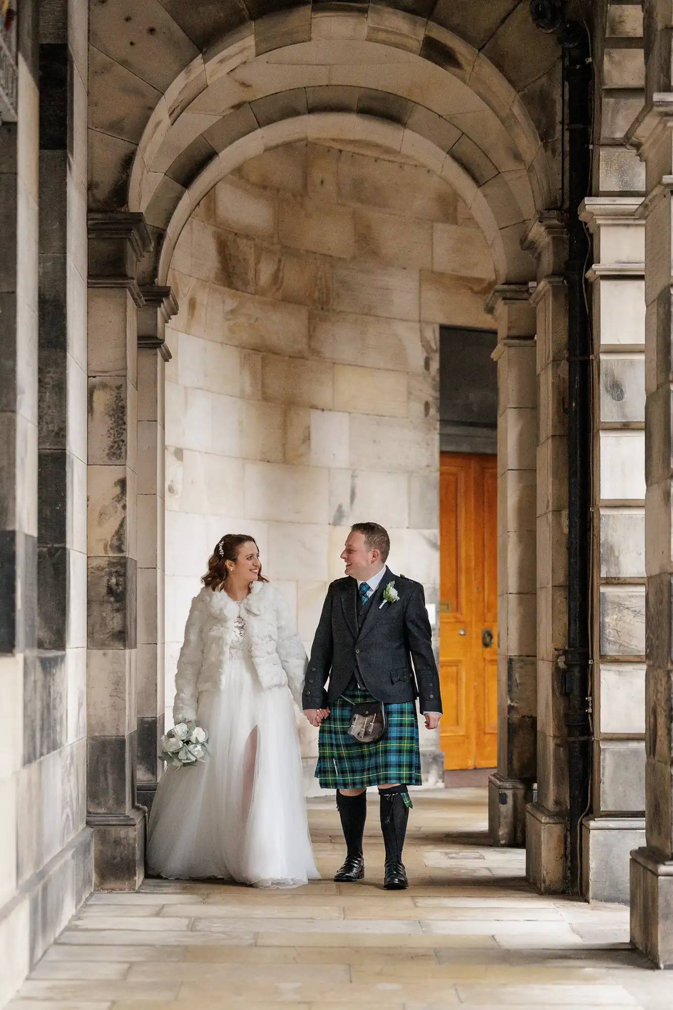 A bride in a white gown and fur jacket holds a bouquet and smiles at a groom in a blue jacket and green tartan kilt. They walk hand in hand under a stone archway with columns, looking at each other happily. Warm sunlight highlights the old architecture.
