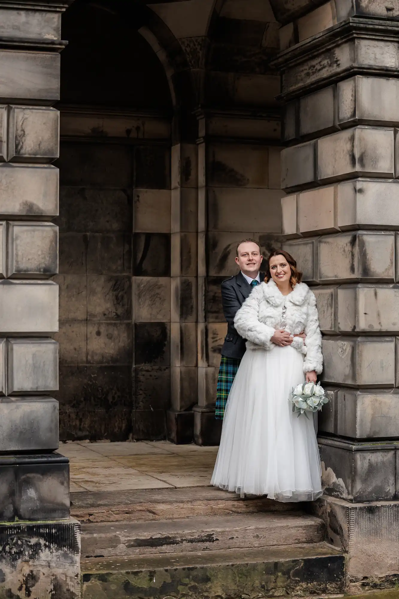 A bride and groom stand smiling under a stone archway. The bride wears a white gown, a white fur jacket, and holds a bouquet of white flowers. The groom stands behind her in a dark suit with a tartan kilt. The setting features large grey stone blocks.