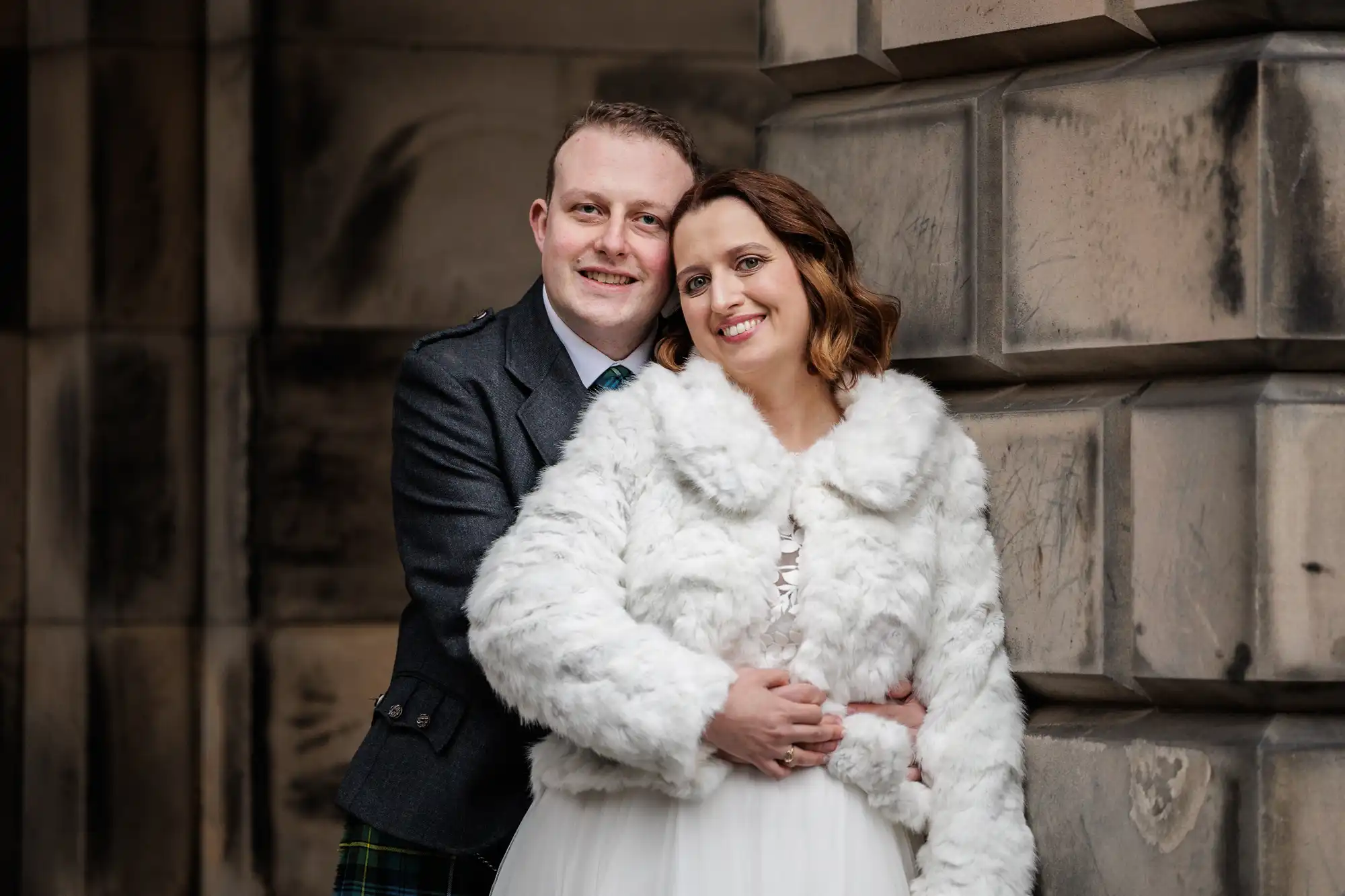 A smiling couple pose closely together. The man wears a dark suit with a kilt, holding the woman from behind. The woman wears a white faux fur jacket over a white dress. They stand against a stone wall, looking happy and warmly dressed.
