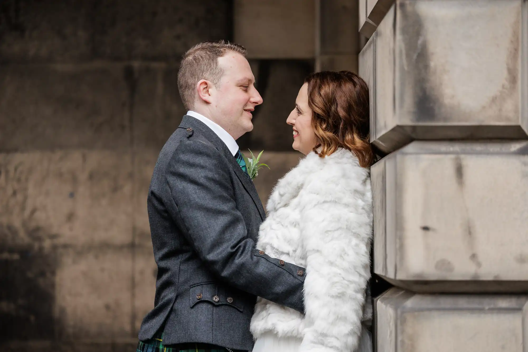 A couple stand close together outdoors by a stone wall. The man, in a dark jacket and kilt, faces the smiling woman who is wearing a white faux fur coat. They gaze into each other's eyes warmly in a candid, affectionate moment.