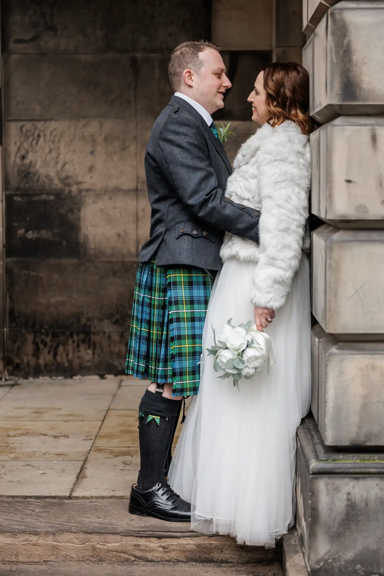 A man in a blue and green tartan kilt and dark jacket lovingly faces a woman in a white furry jacket and long white skirt, holding white flowers. They are standing close together against a stone building, gazing into each other's eyes.