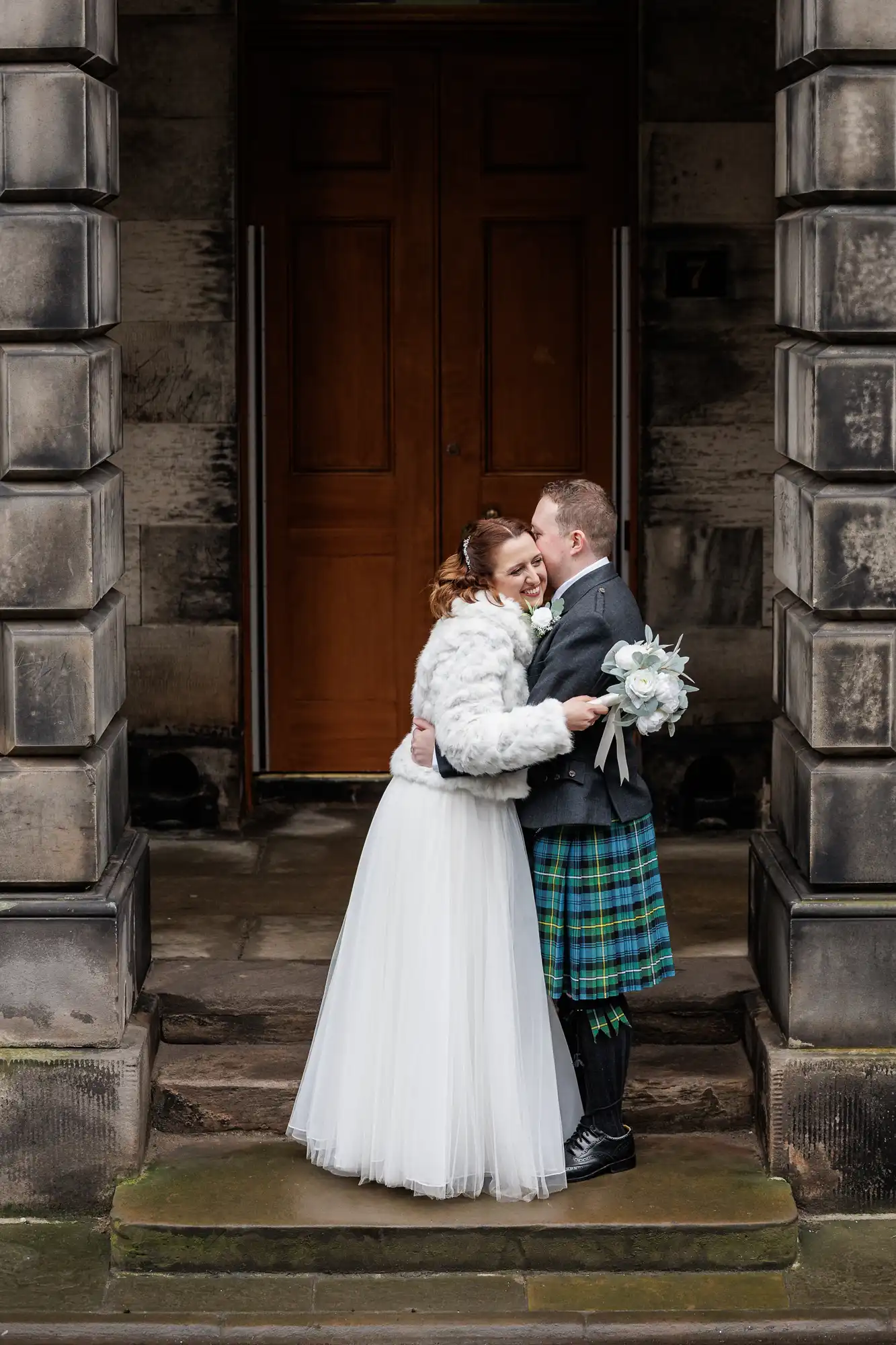A bride in a white gown and fur jacket embraces a groom in a grey jacket and green tartan kilt. They are smiling and standing close on stone steps between two large stone pillars, in front of a wooden door. The scene appears joyful and romantic.