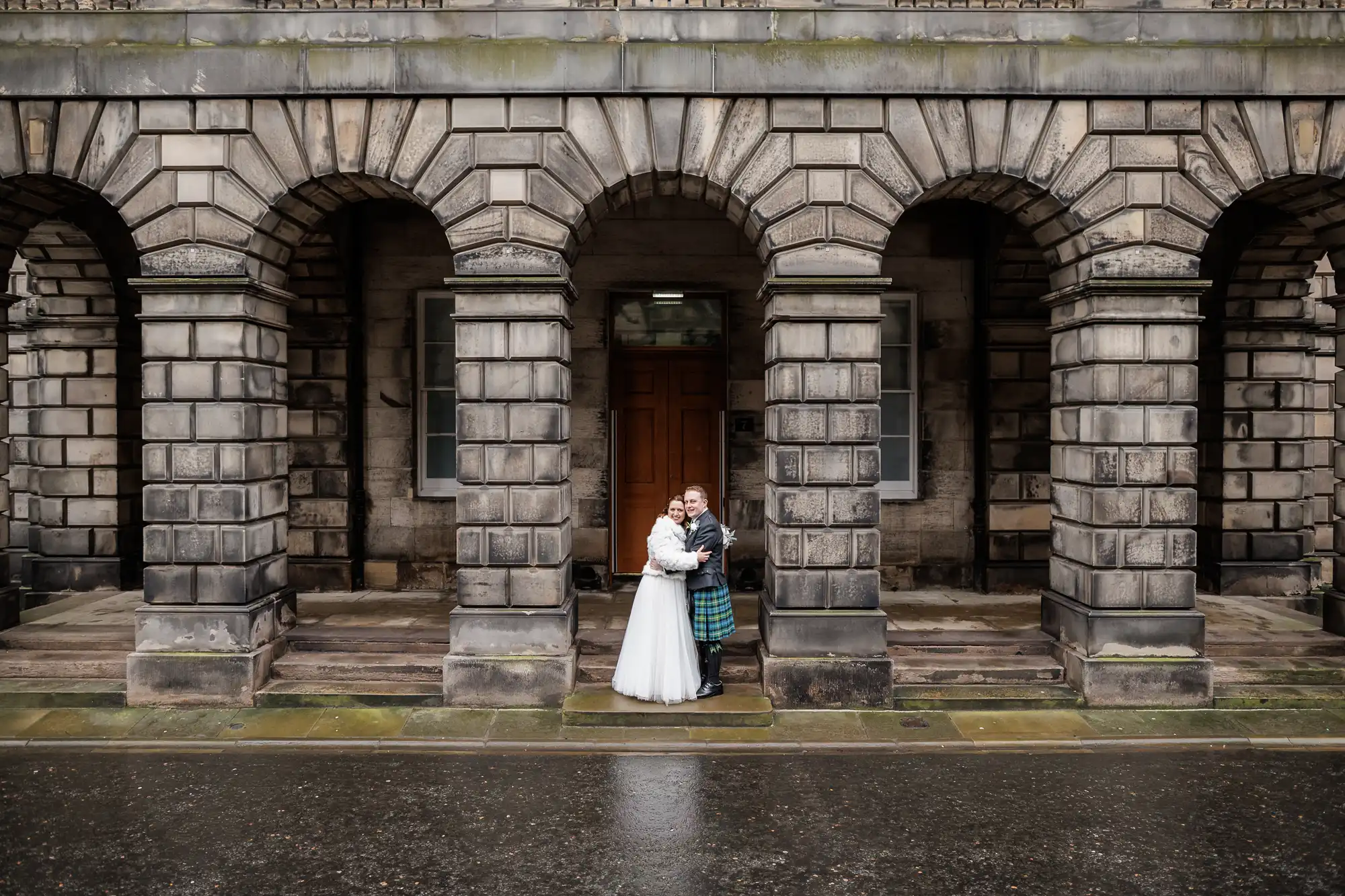 A bride in a white dress and a groom in a kilt stand embracing under stone arches outside a historic, grey stone building on a wet day, centred between three large arched openings, with the ground and stone steps glistening from rain.