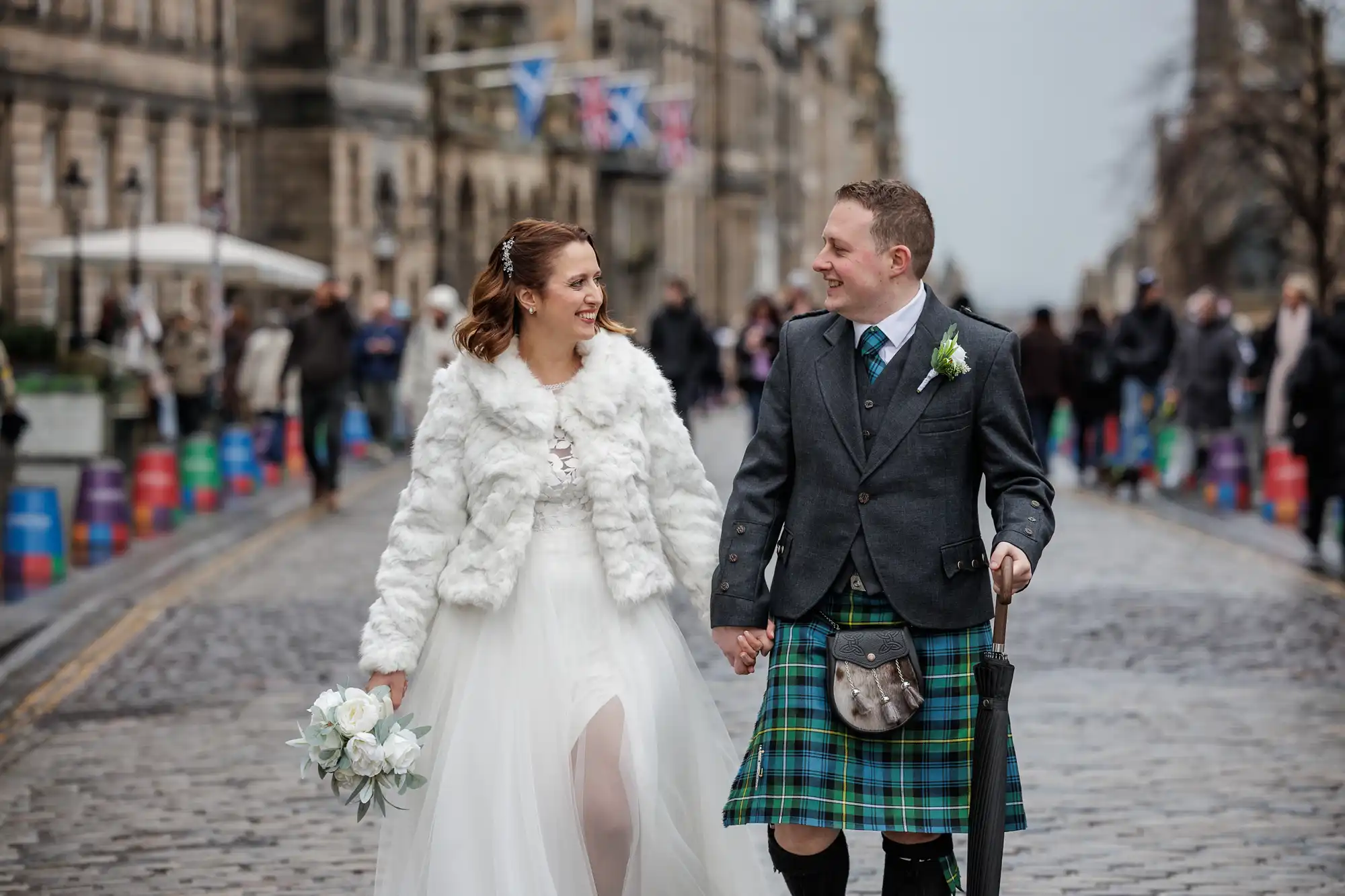 A bride in a white fur jacket and gown holds a bouquet and smiles at a groom wearing a grey jacket, tartan kilt, and sporran. They walk hand-in-hand down a cobbled street lined with blurred people and colourful barrels, with historic buildings in the background.