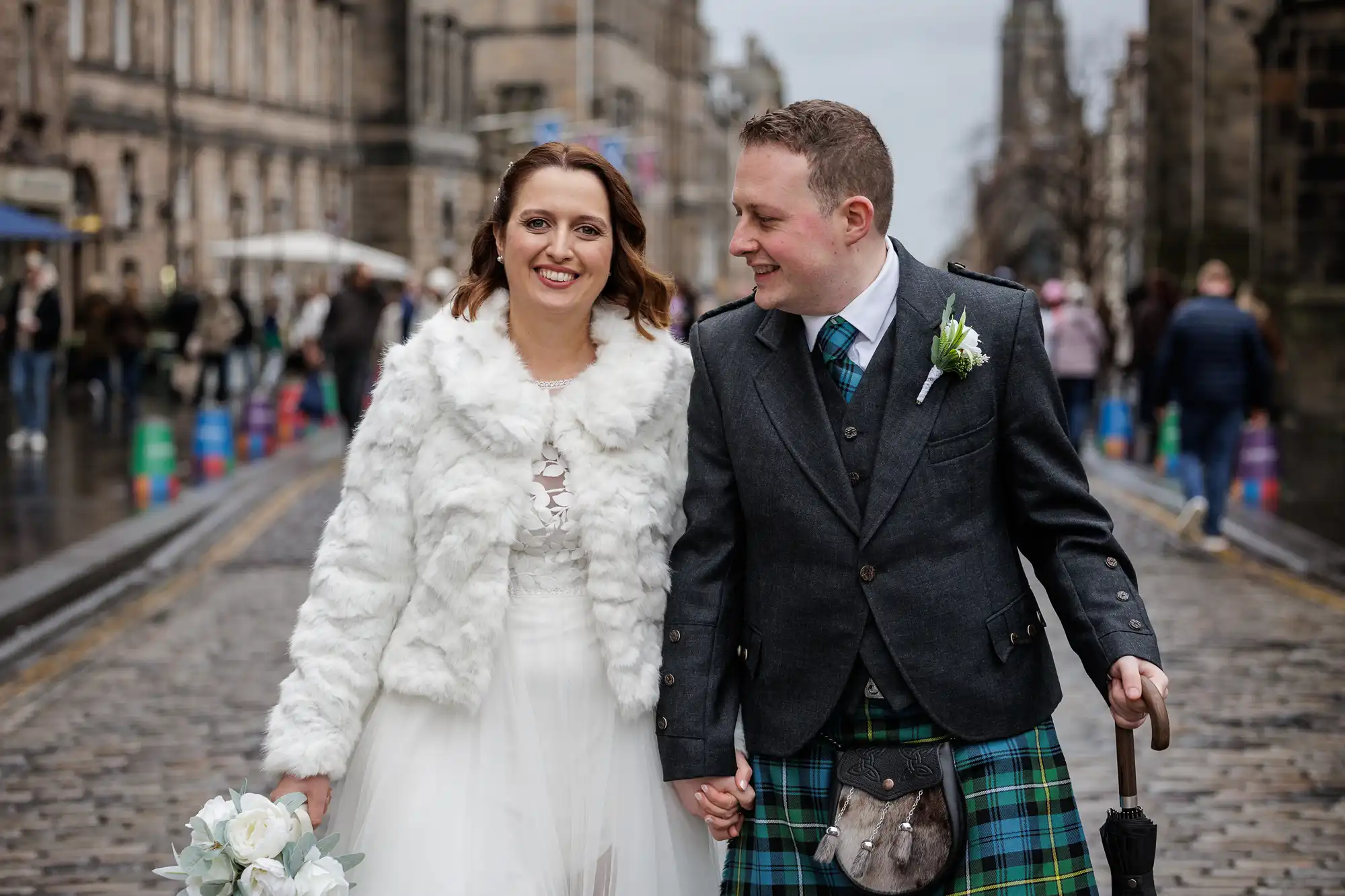 A smiling bride in a white dress and faux fur jacket holds a bouquet and stands beside a groom in a dark jacket, blue tartan kilt, and sporran. They walk hand in hand down a cobbled street with historic stone buildings in the background.