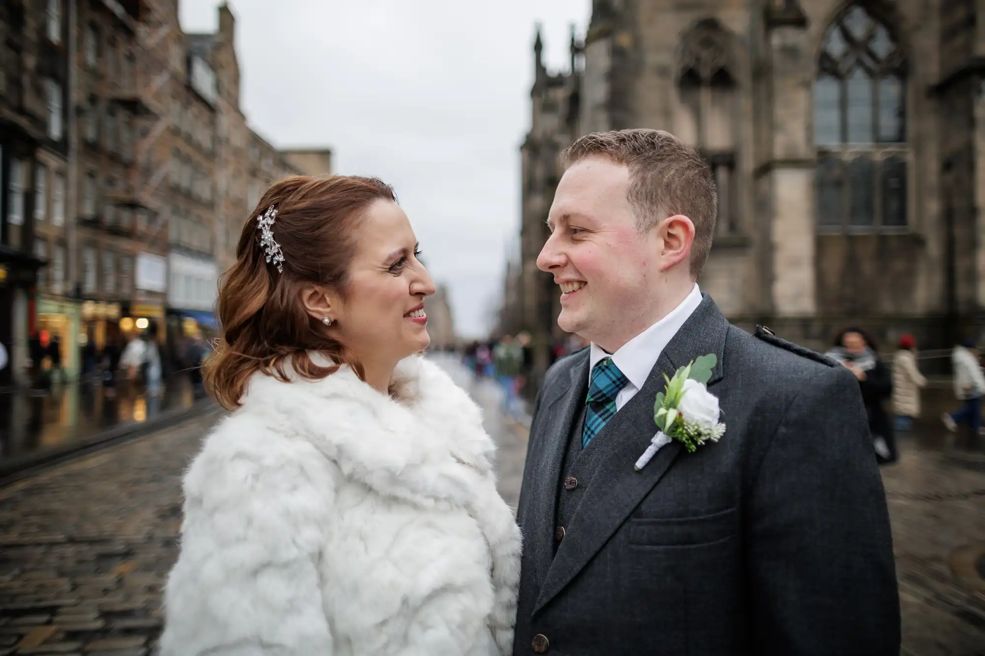 A smiling couple stand close together on a cobbled street. The woman wears a white fur coat with a jewelled hairpin, and the man wears a dark suit with a white buttonhole and checked tie. Gothic buildings and people are visible in the blurred background.