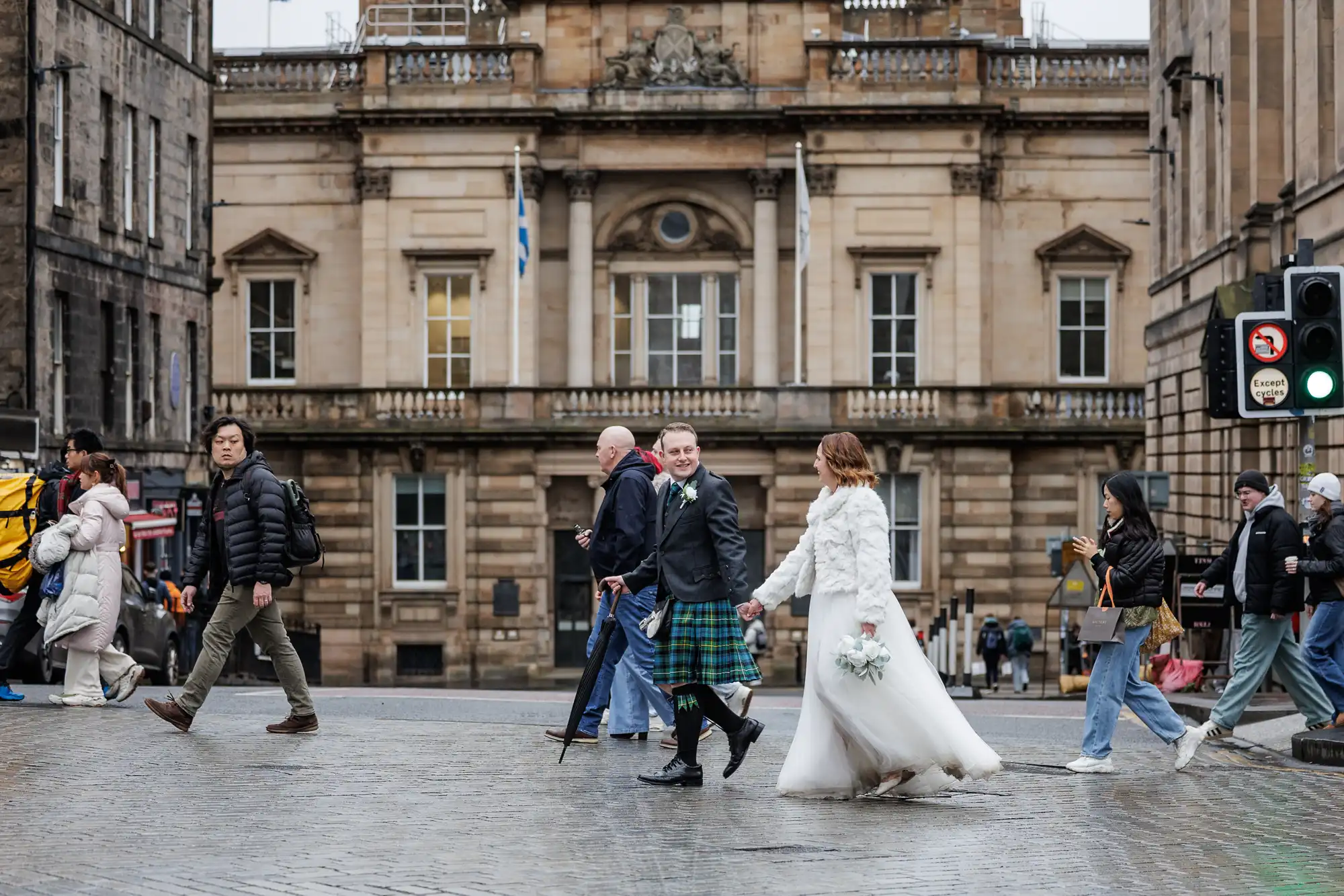A bride in a white dress and a groom in a kilt hold hands whilst crossing a cobbled street. Several pedestrians walk by. Behind them is a large historic stone building with columns and windows. The scene is lively and urban.