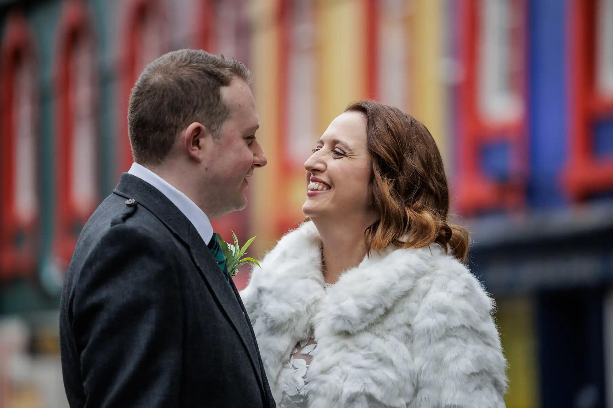 A smiling couple in formal attire faces each other outdoors. The woman wears a white faux fur coat and the man a dark suit with a buttonhole. Colourful, blurred buildings form the background, highlighting their joyful expressions.
