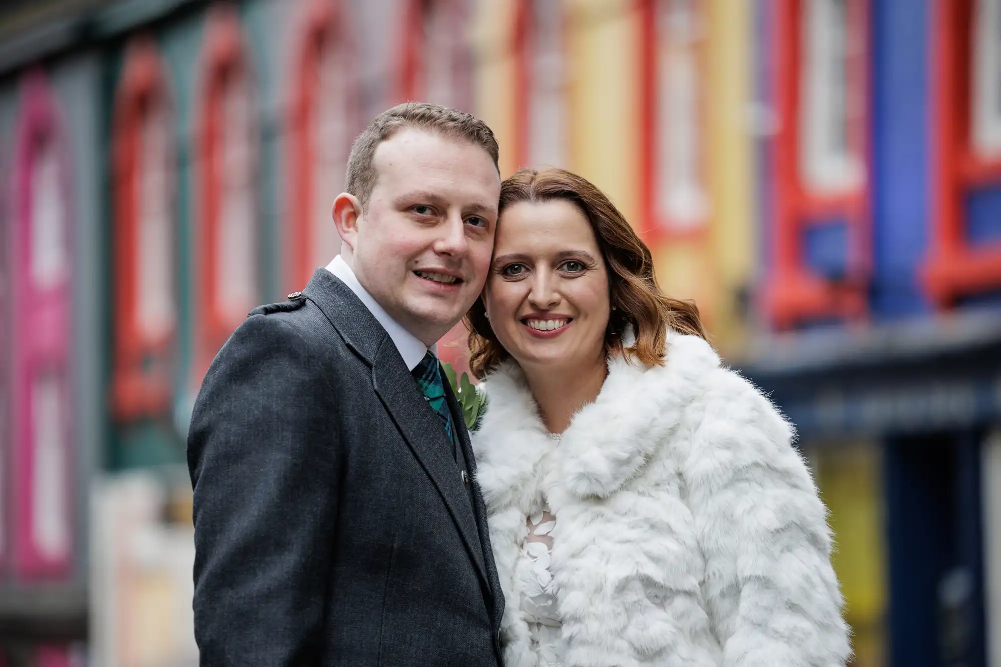 A couple pose closely and smile at the camera. The man wears a dark suit, and the woman wears a white fur coat. They stand on a street lined with colourful buildings in shades of red, blue, yellow, and green, which appear softly blurred in the background.