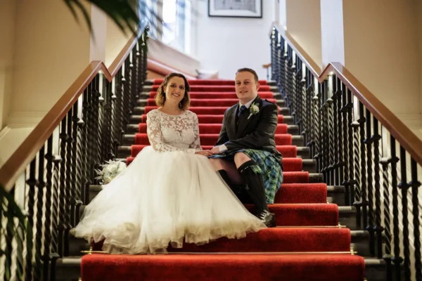 Virgin Hotels Edinburgh wedding photographer image of a bride in a white lace wedding dress and a groom in a blue tartan kilt sit side by side on red-carpeted stairs, holding hands and smiling. The staircase has black railings and white walls, and a bouquet rests beside the bride.