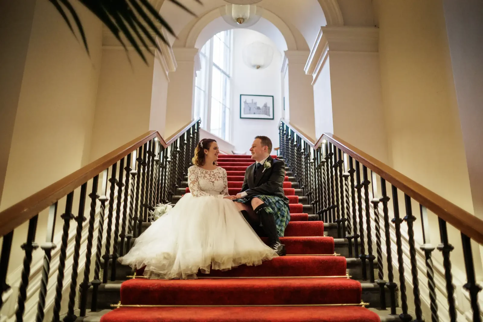 A bride in a white gown and a groom in a dark jacket and kilt sit together on a red-carpeted staircase, smiling at each other in a grand, well-lit interior with high arches and decorative railings.