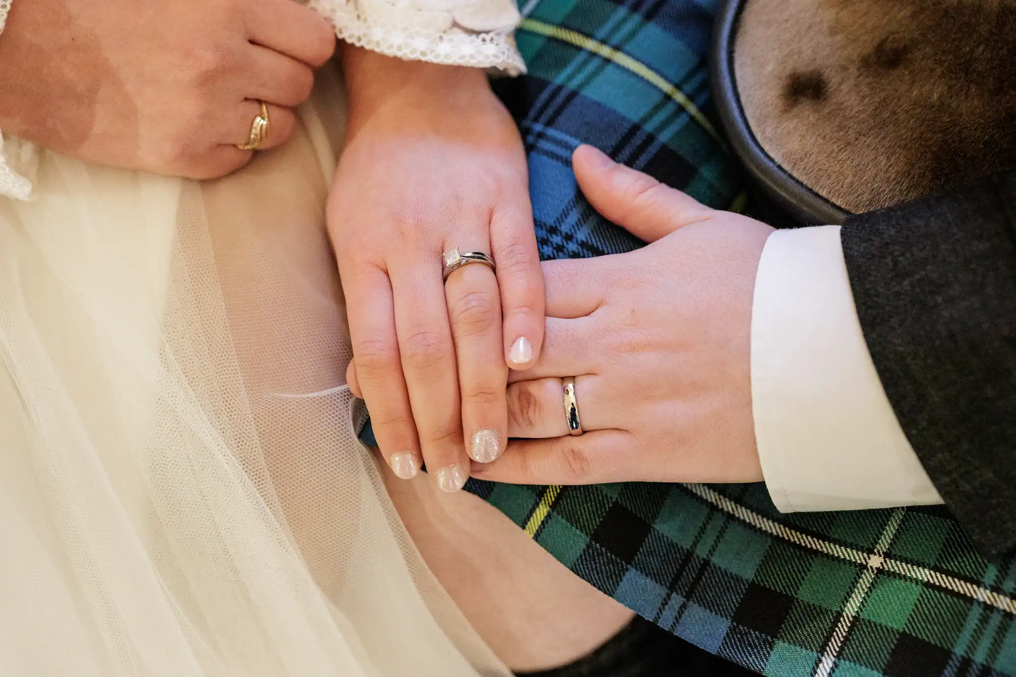 Close-up of two hands with wedding rings resting on top of each other; one hand wears a white dress, the other a dark suit sleeve and blue-green tartan kilt. The scene suggests a wedding or commitment ceremony.