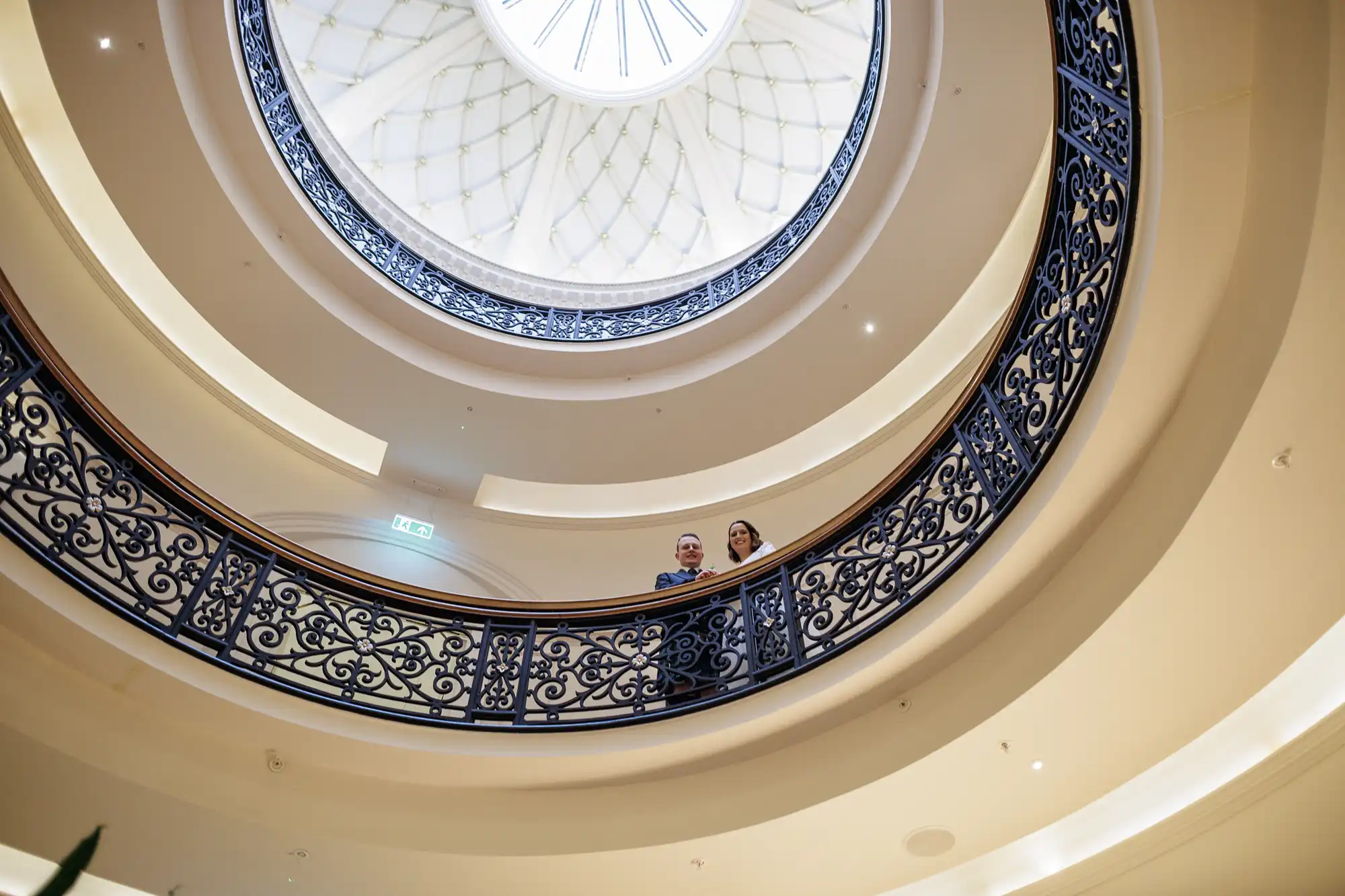 Two people stand on an ornate circular balcony, looking down. The photo is taken from below, capturing decorative railings and a domed glass ceiling above, which lets in natural light. The elegant architecture creates a sense of openness and grandeur.