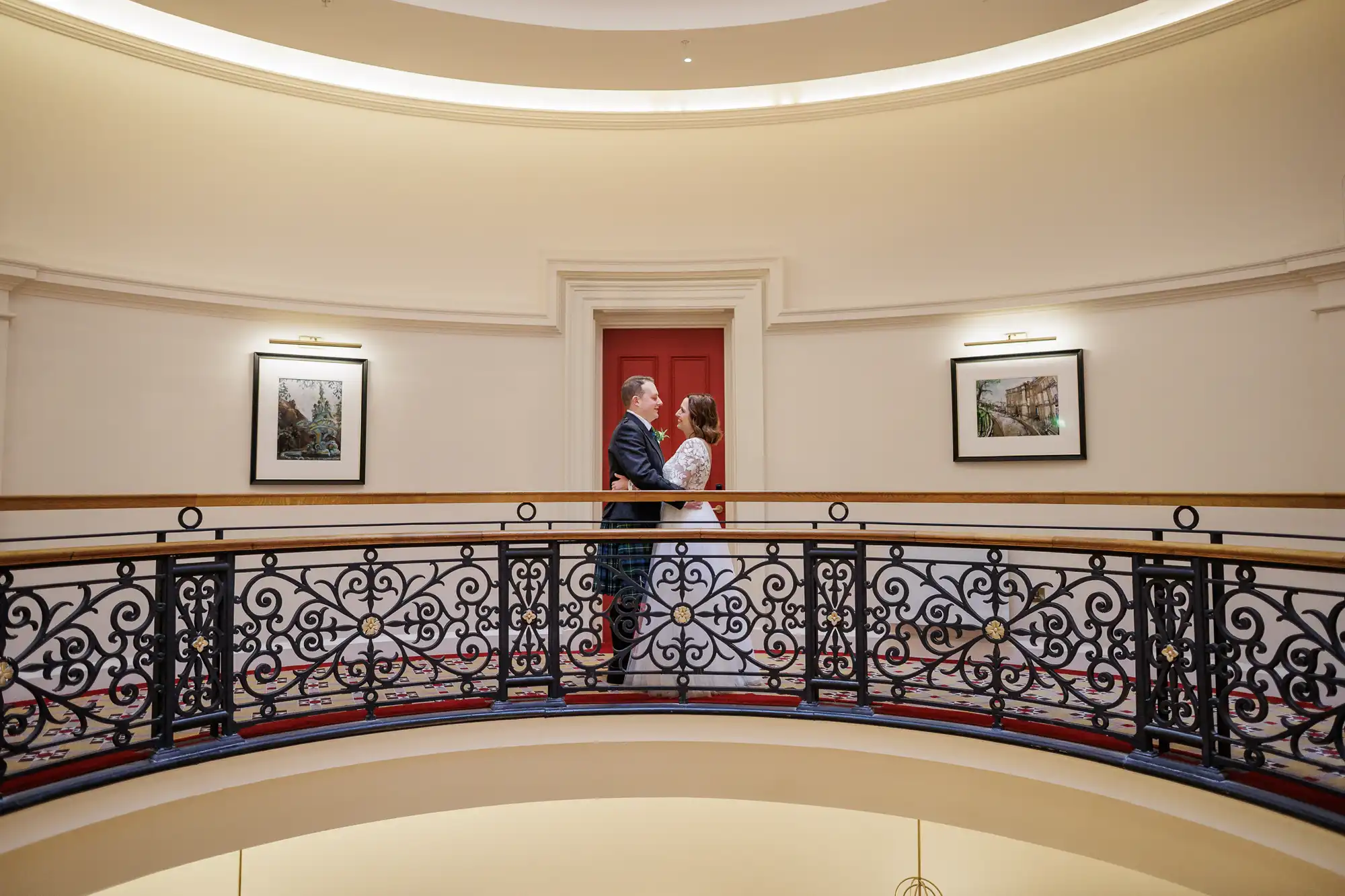 A couple in formal attire stands closely together on an ornate, curved balcony indoors. The background features cream walls, framed artwork, and a red door centred behind them. Warm lighting creates a soft ambience in the elegant setting.