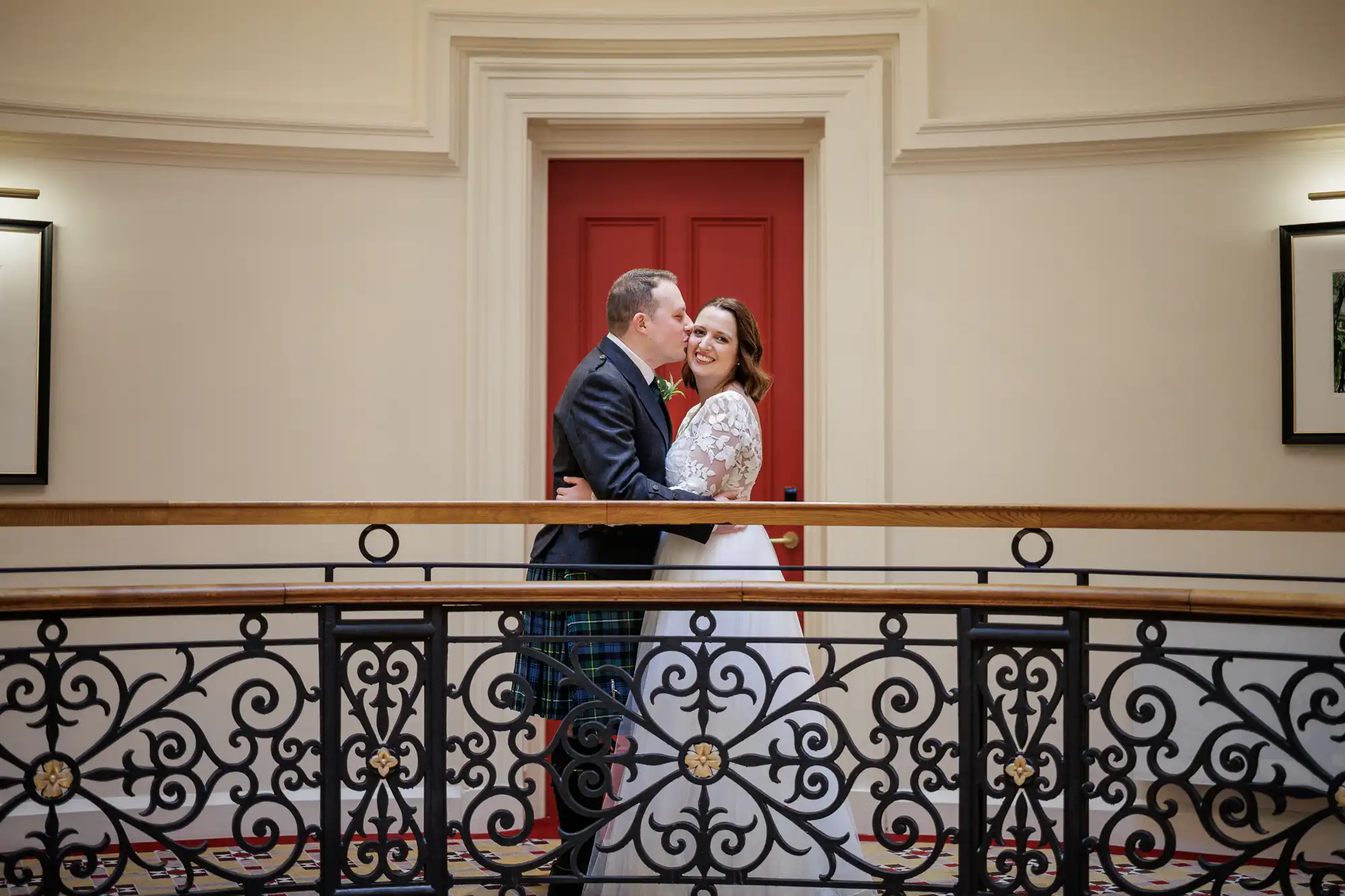 A bride in a white dress and a groom in a dark suit and kilt embrace and smile at each other on an ornate balcony with black and gold railings, in front of a red door with cream-coloured walls. Framed pictures hang on both sides.