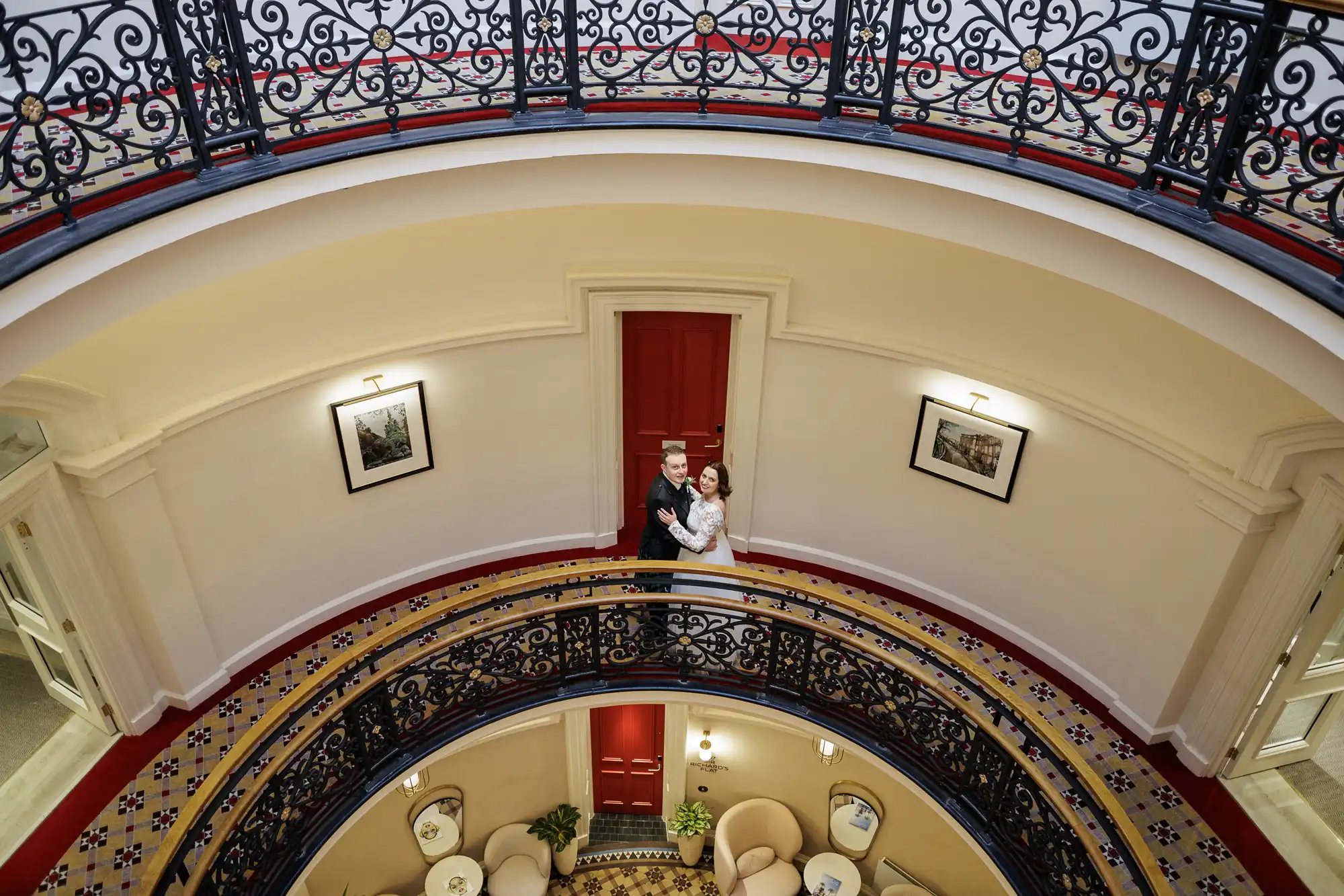 A bride and groom stand by an ornate black railing on a circular upper balcony inside a grand building. A red door is behind them, with framed pictures on cream-coloured walls and patterned tiles on the floor below. Lounge chairs are visible on the lower level.