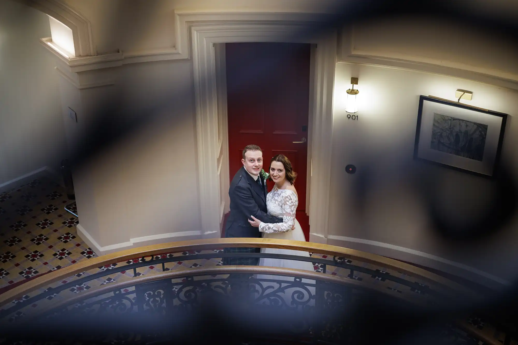 A couple in formal attire stands embracing at the bottom of a staircase with ornate bannisters. The woman wears a white dress and the man a dark suit. They look up towards the camera. The corridor is softly lit, with patterned floor tiles and framed art on the wall.