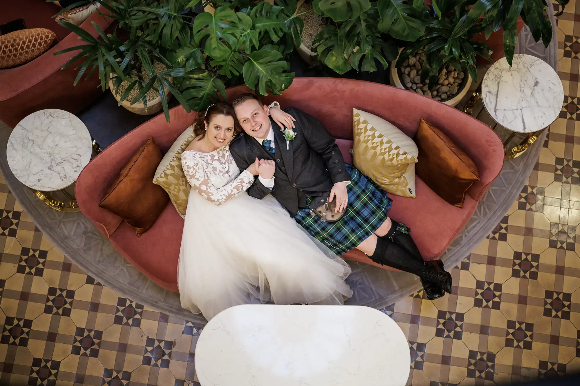 A bride in a white dress and a groom in a kilt sit on a curved pink sofa, surrounded by green plants and patterned floor tiles. They smile up at the camera, holding hands, with white marble tables and gold cushions nearby.