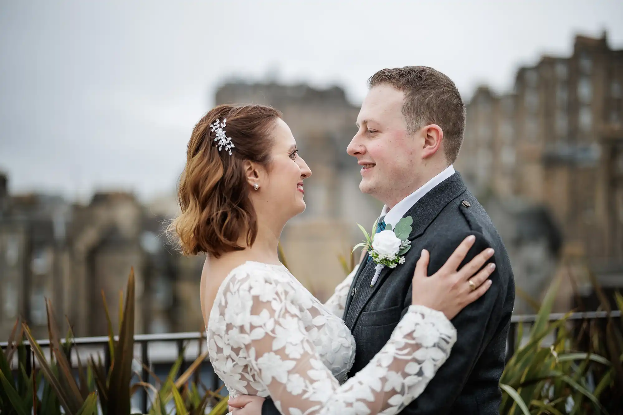 A bride in a white floral lace dress and a groom in a dark suit with a buttonhole smile at each other, embracing outdoors with blurred historic buildings and plants in the background, under a cloudy sky.