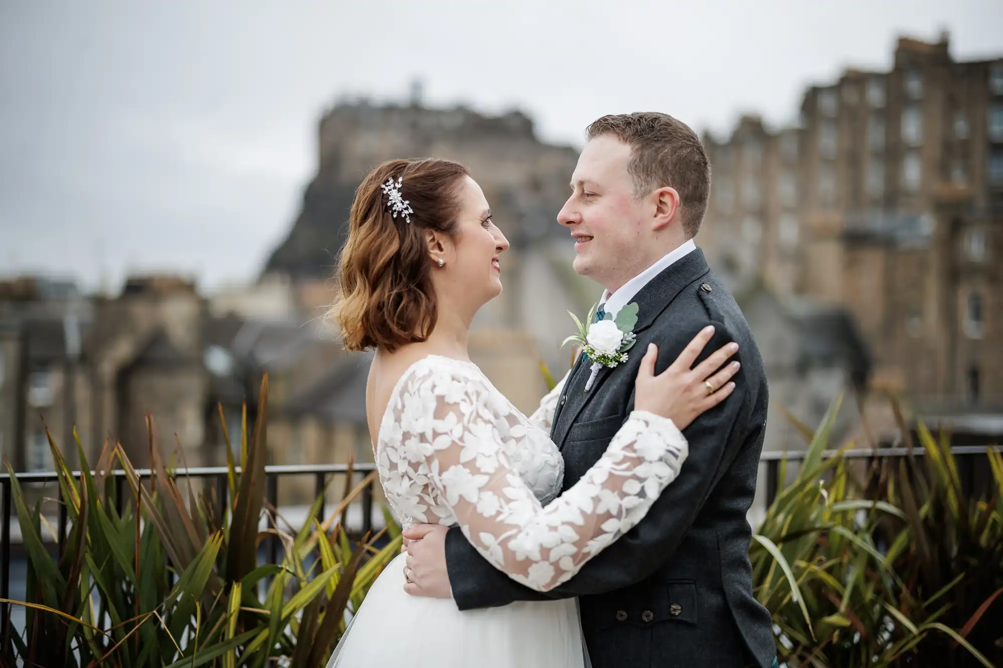 A bride and groom embrace outdoors, smiling at each other. The bride wears a white dress with floral lace sleeves and a hair accessory. The groom wears a dark suit with a buttonhole. Old stone buildings and a castle are visible in the blurred background.