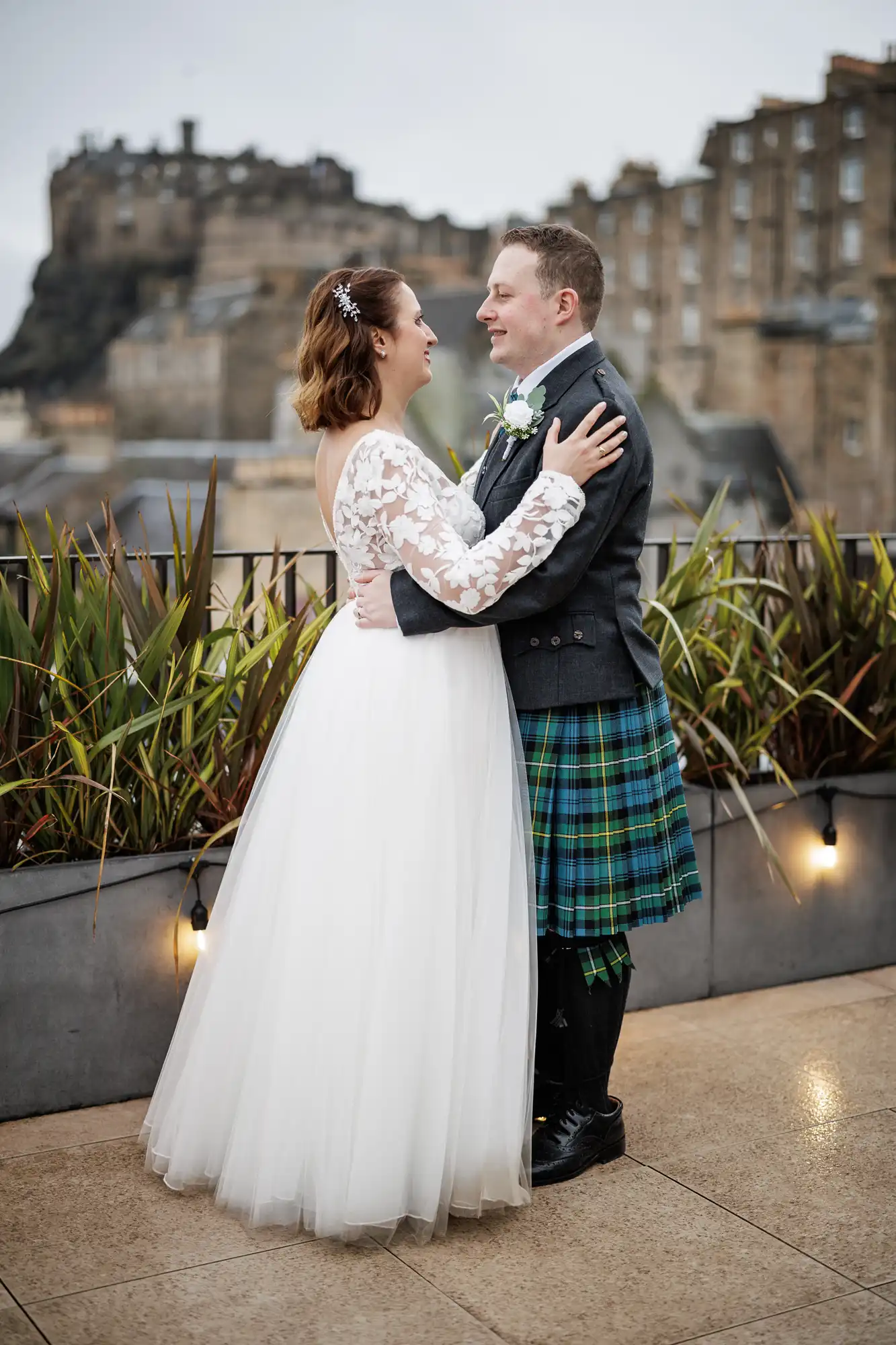 A bride in a white dress and a groom in a Scottish kilt stand facing each other, embracing and smiling on a rooftop with potted plants. Historic stone buildings and a large castle are visible in the background on an overcast day.
