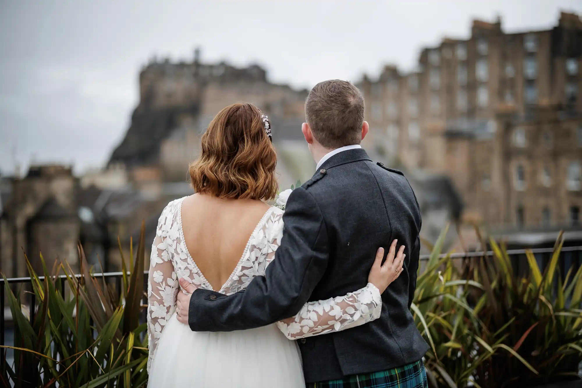 A bride in a white dress with lace sleeves and an open back stands next to a groom in a dark jacket and tartan kilt. They face away, arms round each other, looking towards an old stone castle and historic buildings under a cloudy sky.