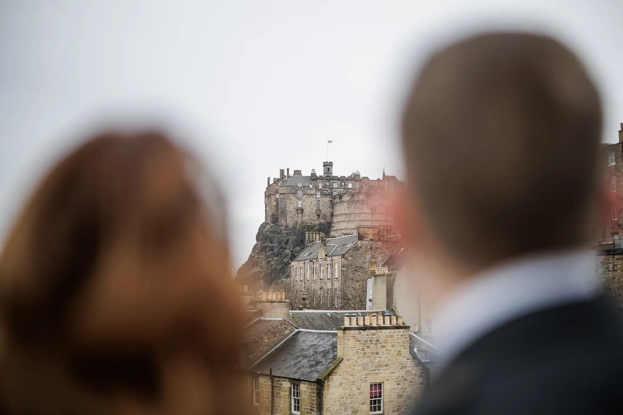A blurred man and woman in the foreground look towards Edinburgh Castle, which stands on a rocky hill under an overcast sky. The historic stone fortress dominates the background, with slate-roofed buildings below.