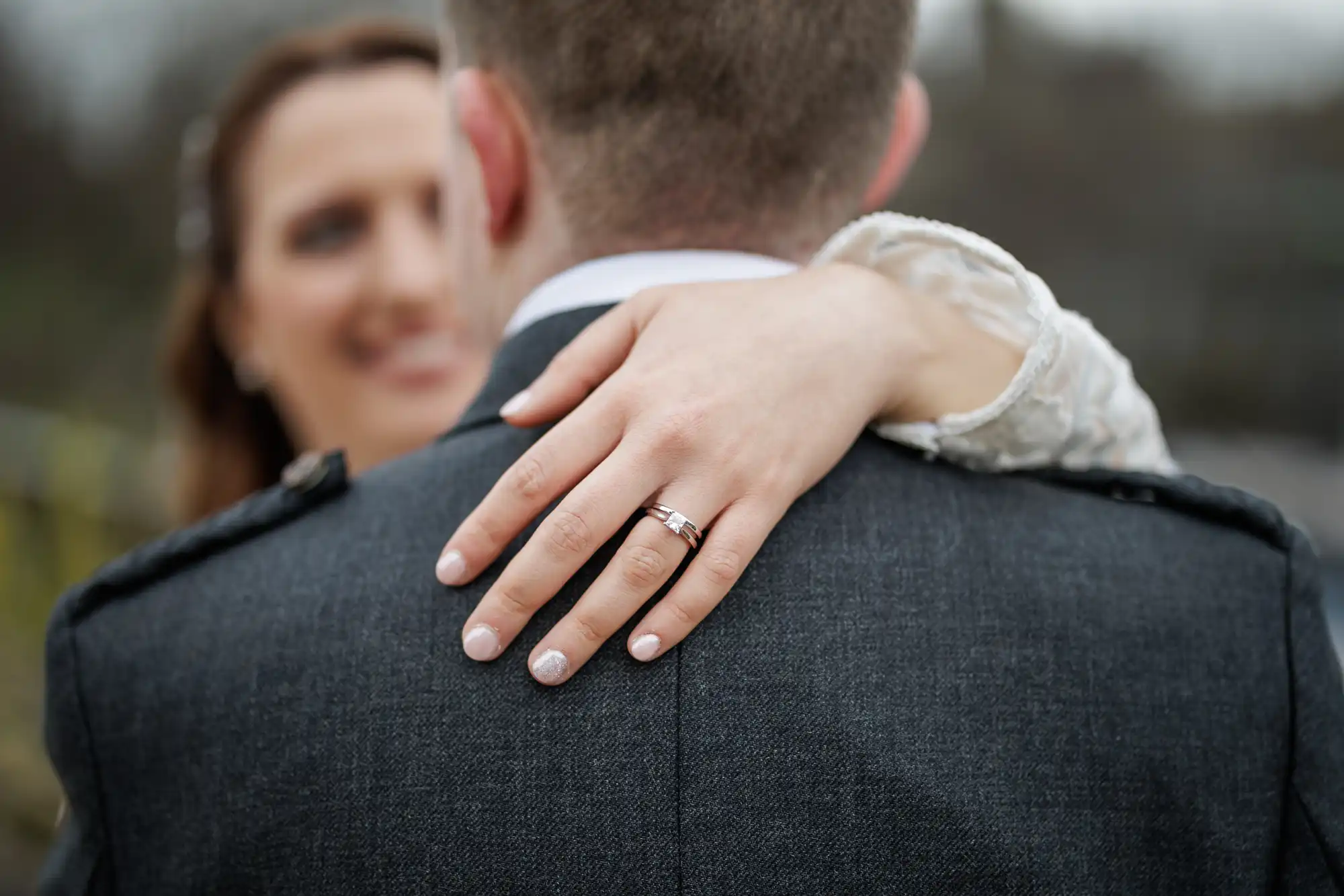 A woman in a white dress embraces a man in a dark suit from behind, showing her hand with a wedding ring on his shoulder. The background is blurred, focussing on her ring and hand. Both appear happy and possibly newly married.