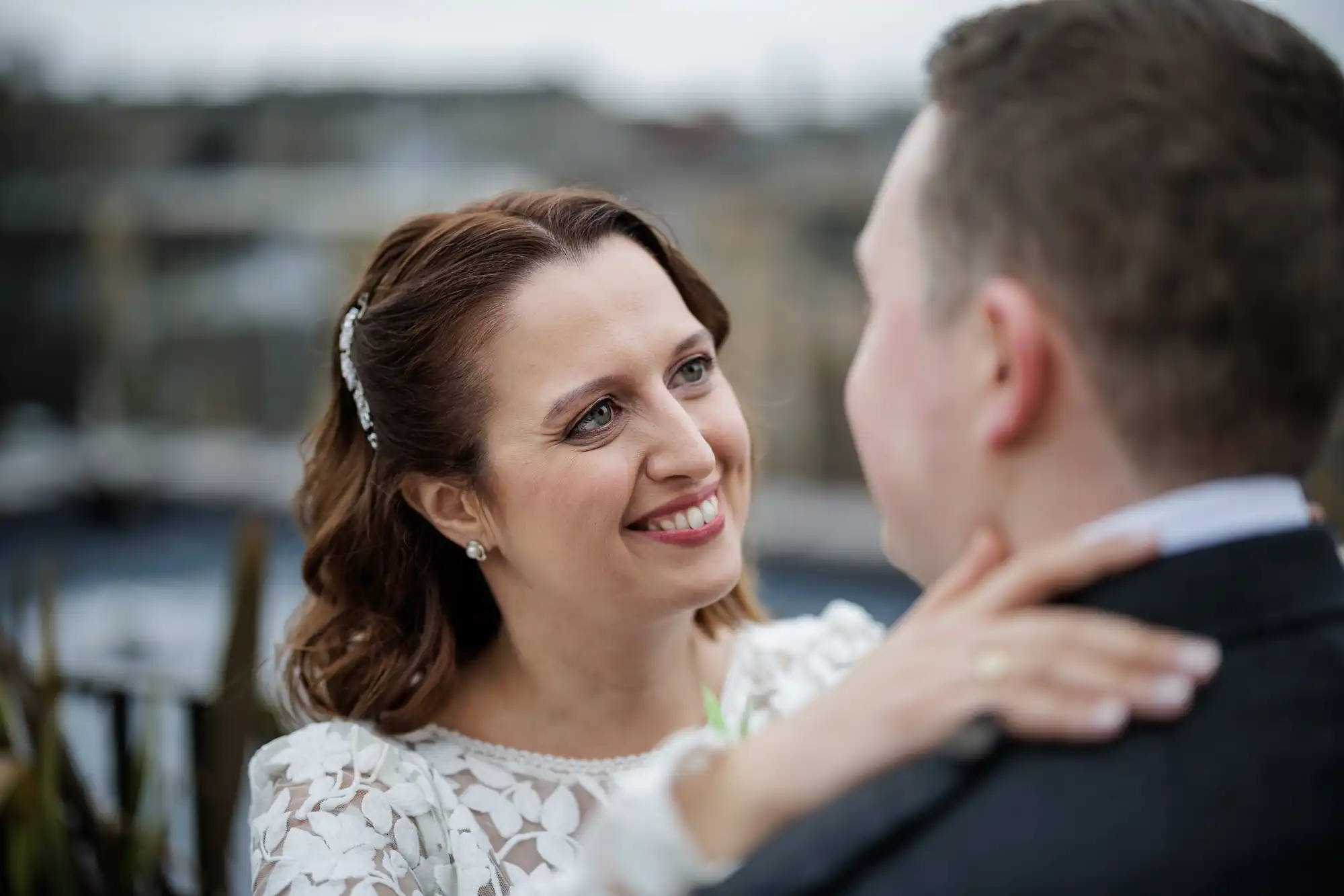 A woman in a white lace bridal dress smiles warmly at a man, gently holding his neck. The man’s face is turned away from the camera. The background is softly blurred, suggesting an outdoor setting. The woman’s hair is styled and she wears small earrings.