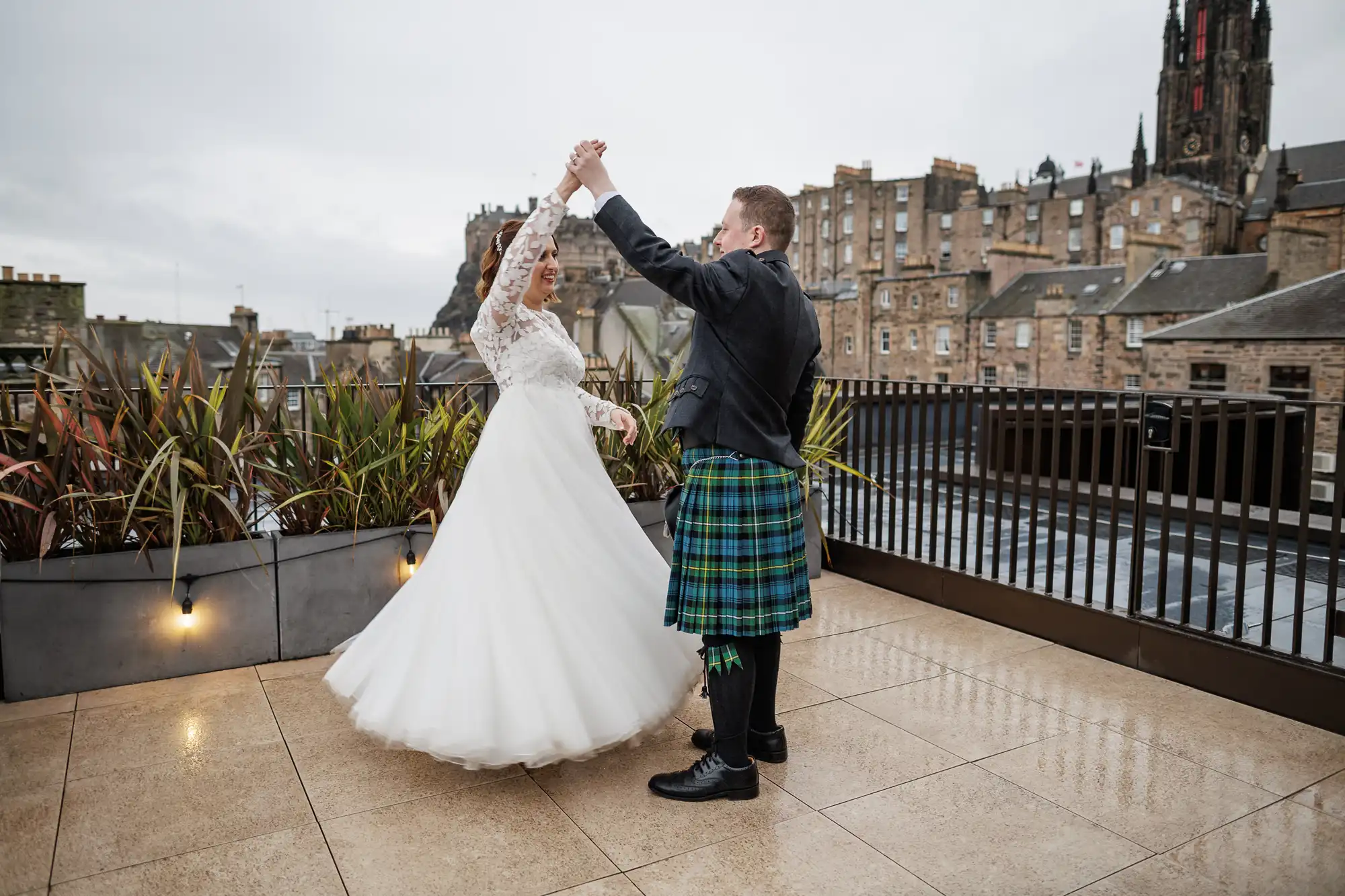 A bride in a white dress and a groom in a traditional Scottish kilt dance on a rooftop terrace with city buildings and a church spire in the background. The sky is overcast, and plants line the terrace, creating a romantic atmosphere.