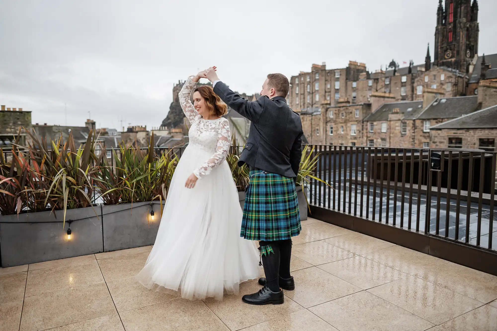 A bride in a long white gown twirls on a rainy rooftop, led by a groom in a traditional Scottish kilt and jacket. Behind them are stone buildings and a church spire, under a cloudy sky. Green plants line the terrace, and the ground appears wet.