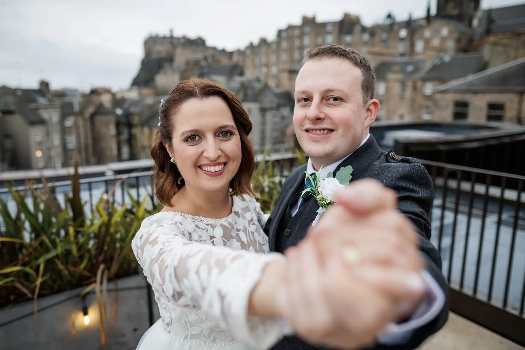 A smiling bride and groom in wedding attire hold hands and pose for a close-up photo outdoors, with historic stone buildings and Edinburgh Castle blurred in the background under a cloudy sky.
