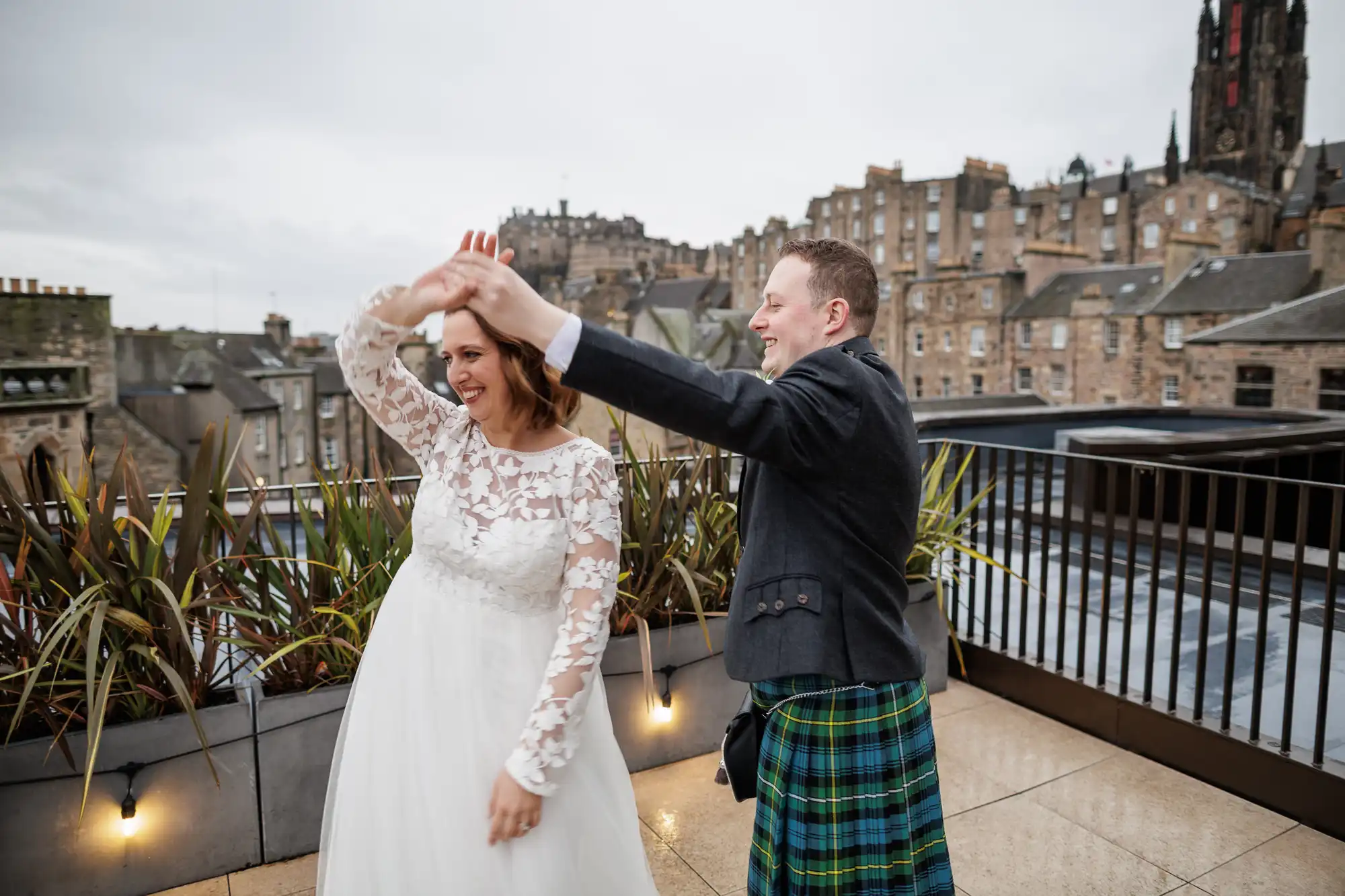 A smiling bride in a white lace wedding dress dances with a groom in a dark jacket and green tartan kilt on a rooftop terrace, with stone buildings and a church spire in the background under an overcast sky.