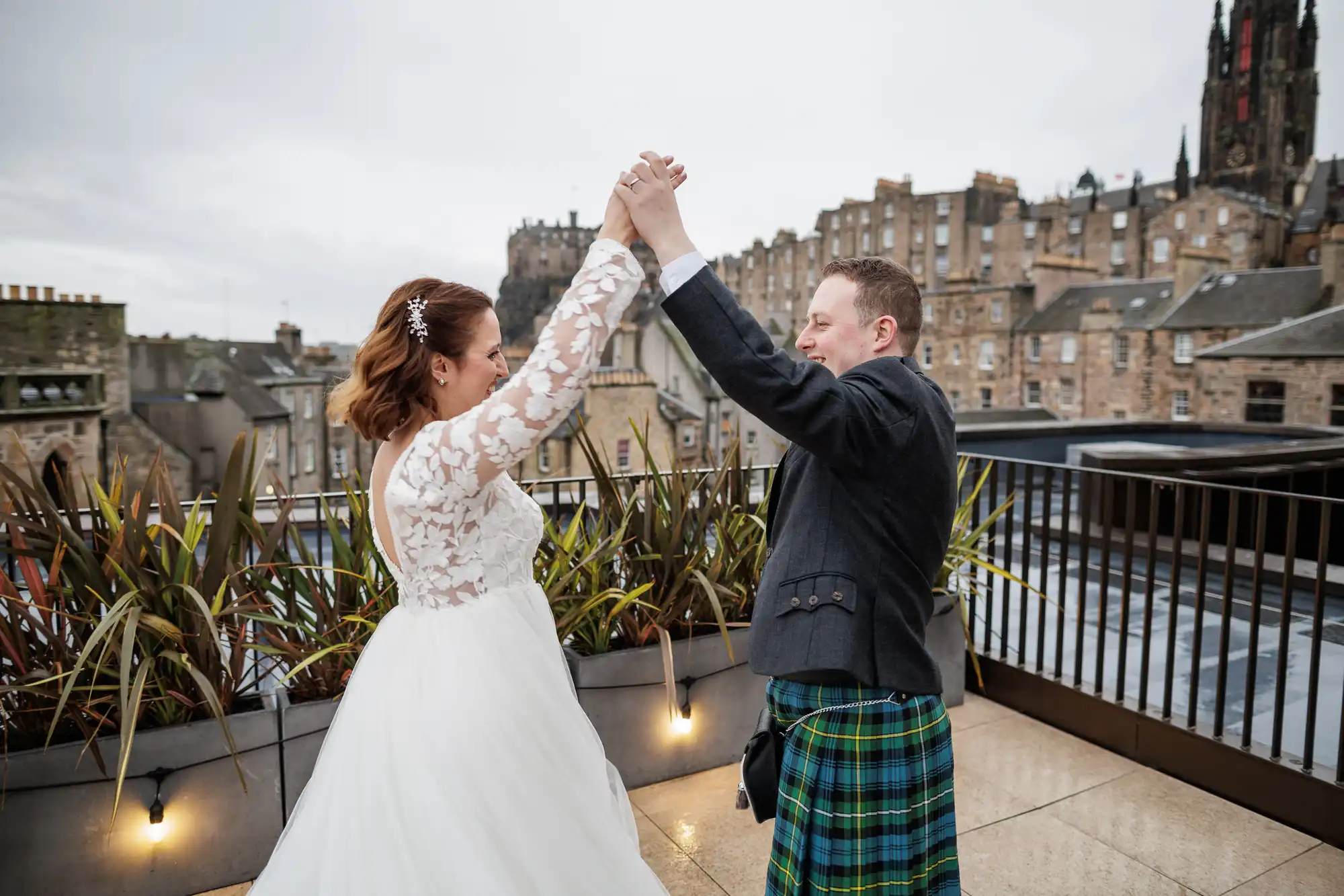 A bride in a white lace-sleeved gown and a groom in a traditional Scottish kilt hold hands and dance on a rooftop terrace. Behind them are city buildings and cloudy skies, with plants lining the edge of the terrace.