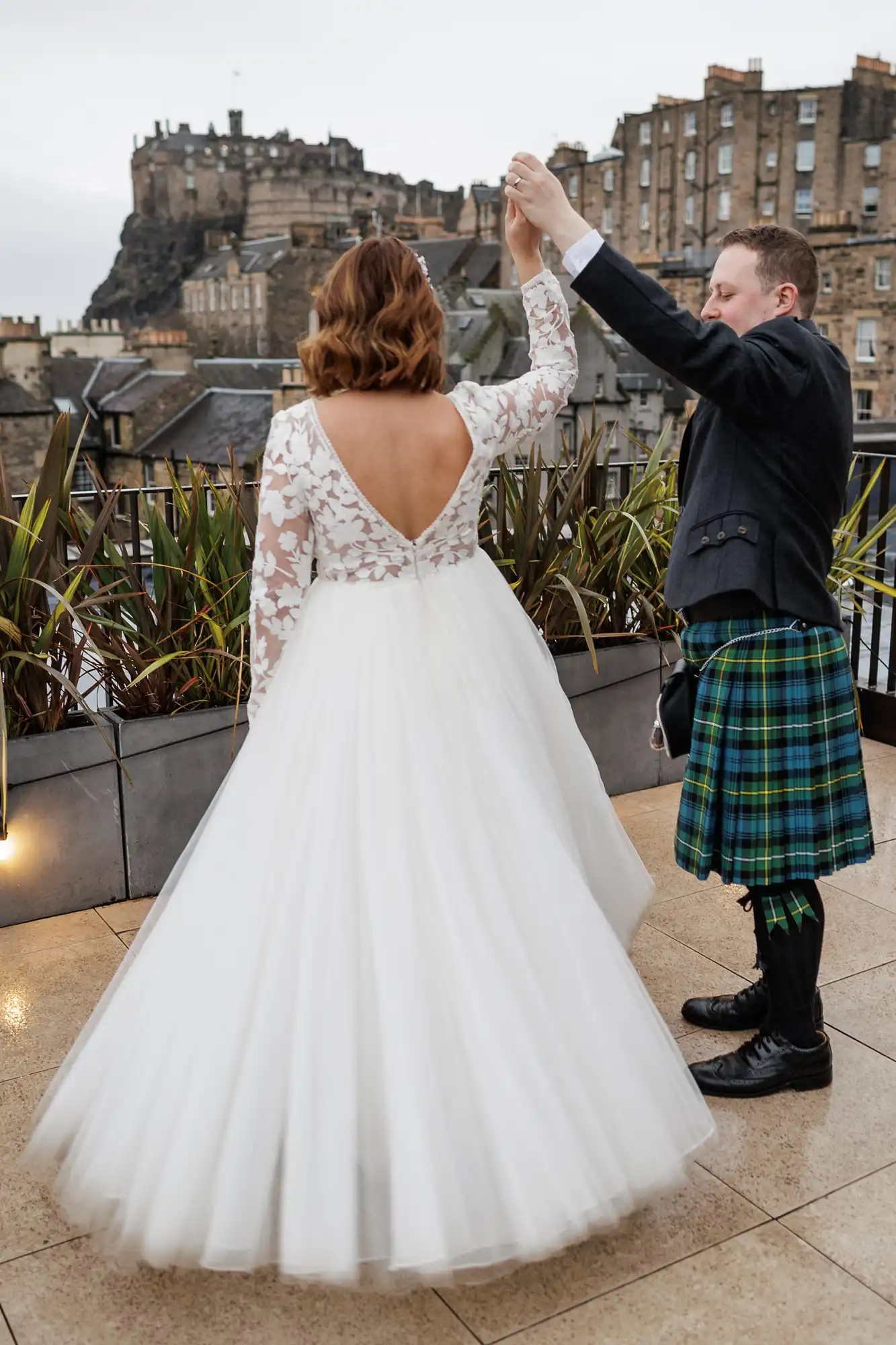 A groom in a green tartan kilt twirls a bride in a white, long-sleeved, open-back lace gown on a rooftop. In the background, historic stone buildings and Edinburgh Castle are visible under a cloudy sky.