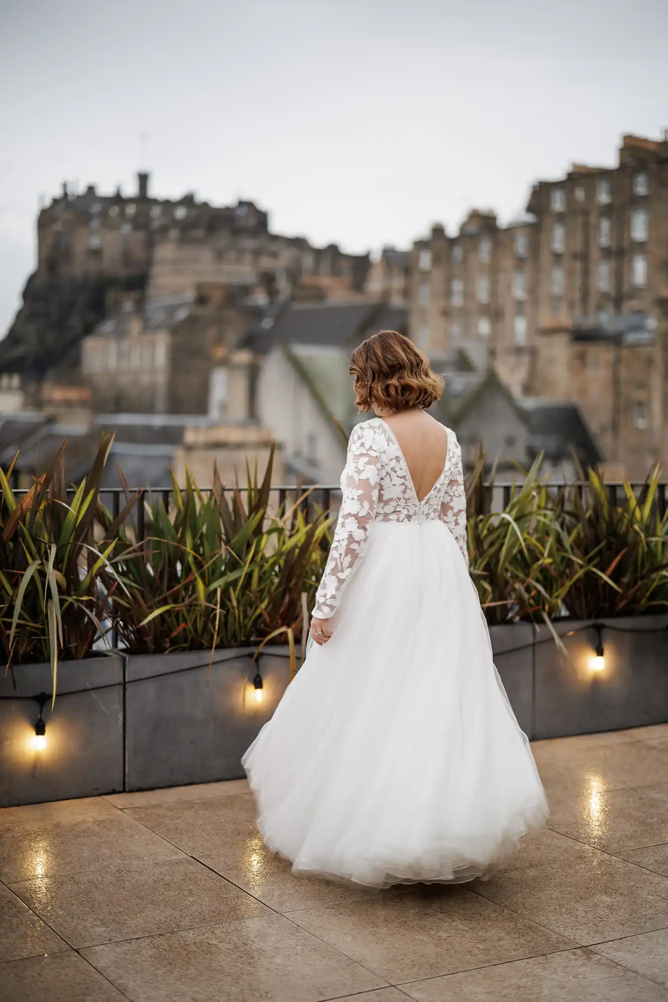 A woman in a long-sleeved white wedding dress with lace details and a deep V back stands on a terrace, facing away. Potted plants line the railing, and historic stone buildings and a castle are visible in the background under a cloudy sky.