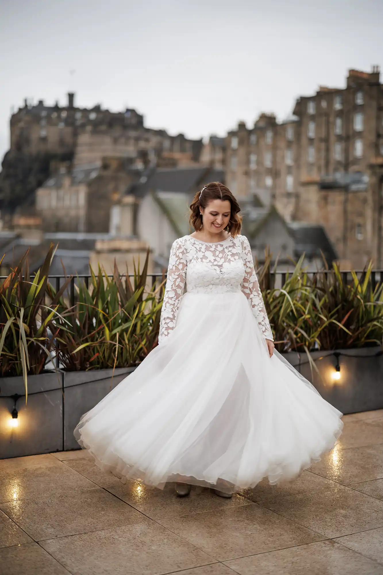 A smiling woman in a long white wedding dress with lace sleeves spins joyfully on a wet rooftop terrace, surrounded by plants. In the background, there are old stone buildings and a distant castle under an overcast sky.