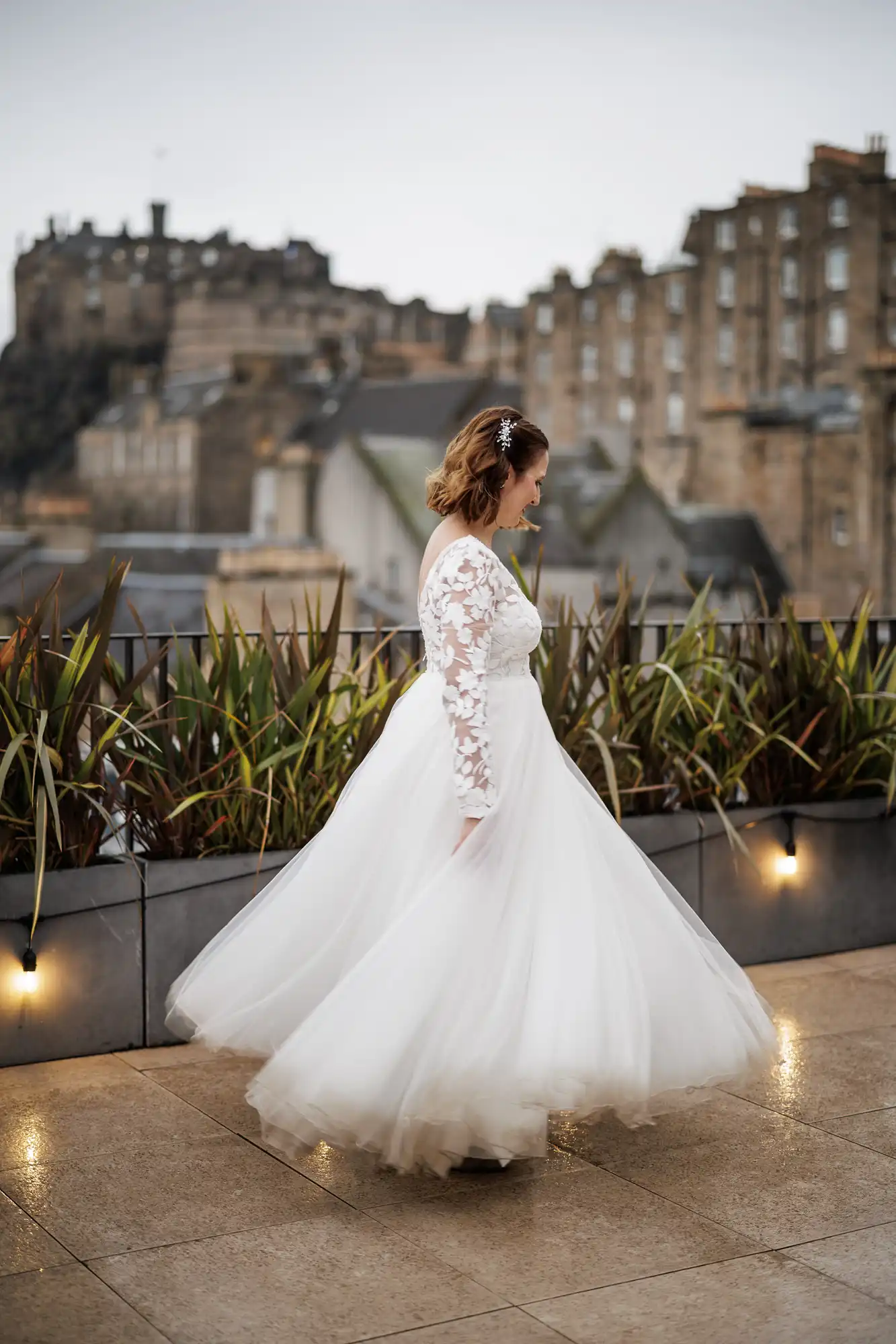A bride in a long-sleeved lace wedding dress twirls on a terrace, her skirt billowing. She has shoulder-length hair and a hair clip. Behind her are green plants, fairy lights, historic stone buildings, and a castle on a hill under a cloudy sky.