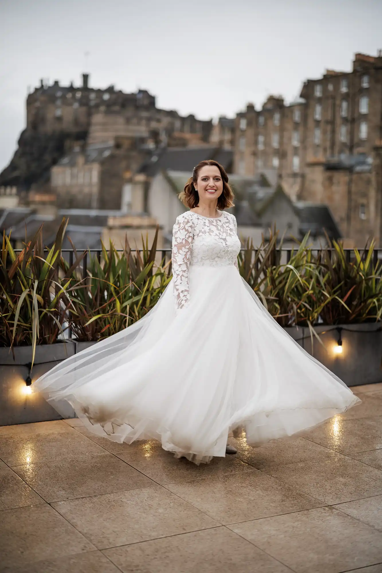 A smiling woman in a long-sleeved, lace wedding dress twirls on a rooftop terrace. Her dress flows out around her. Behind her, there are green plants and old stone buildings, including a castle on a hill under an overcast sky.