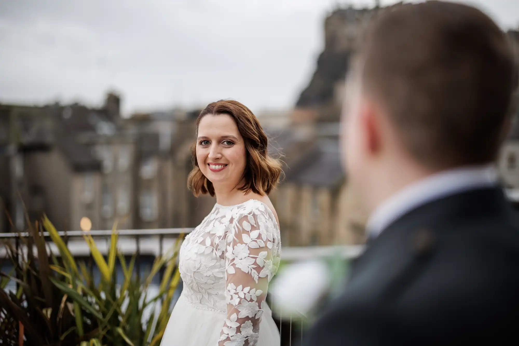 A bride in a white dress with floral lace sleeves smiles at the camera on a balcony with greenery. A blurred person in formal attire is in the foreground. The background shows buildings and a rocky hill under a cloudy sky.