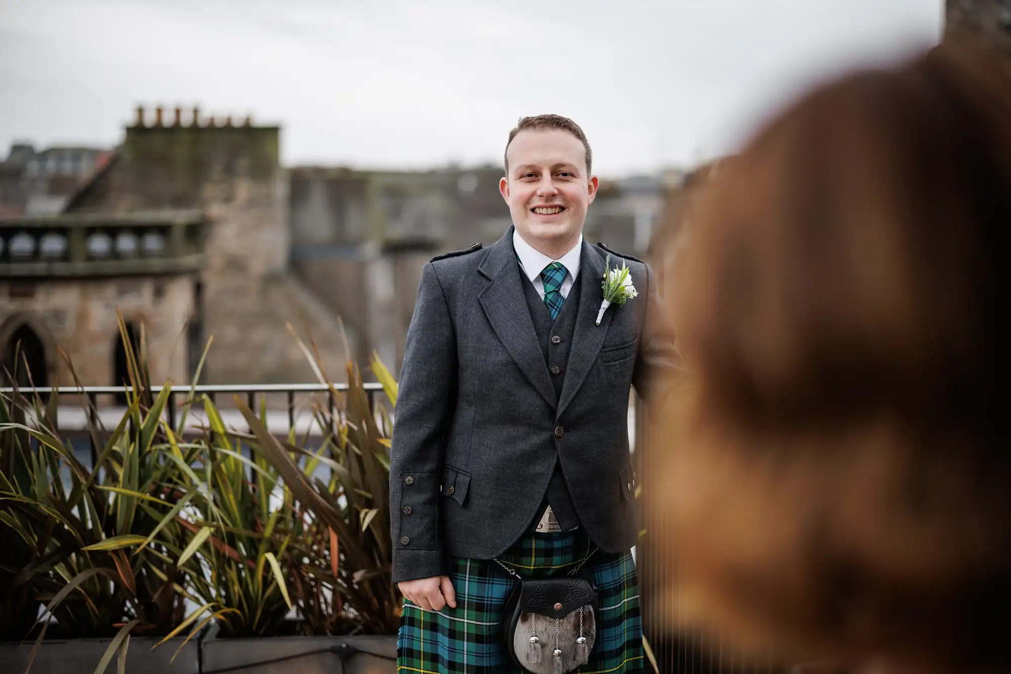 A smiling man in a grey jacket, waistcoat, and green tartan kilt stands outdoors near a railing with plants. He wears a white shirt, tie, and buttonhole. Out of focus in the foreground is the back of someone’s head. Buildings appear in the background.