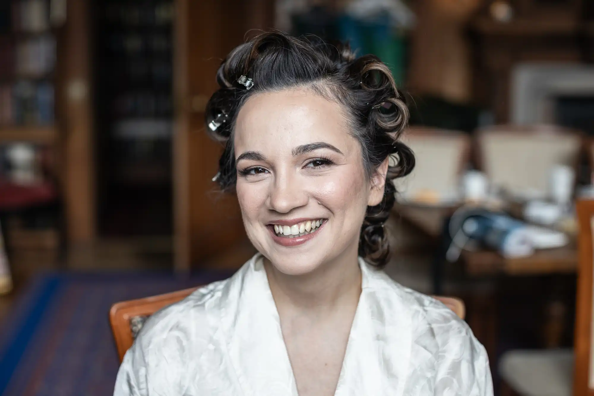 Person smiling, wearing a robe with hair in curlers, seated indoors with a blurred background.