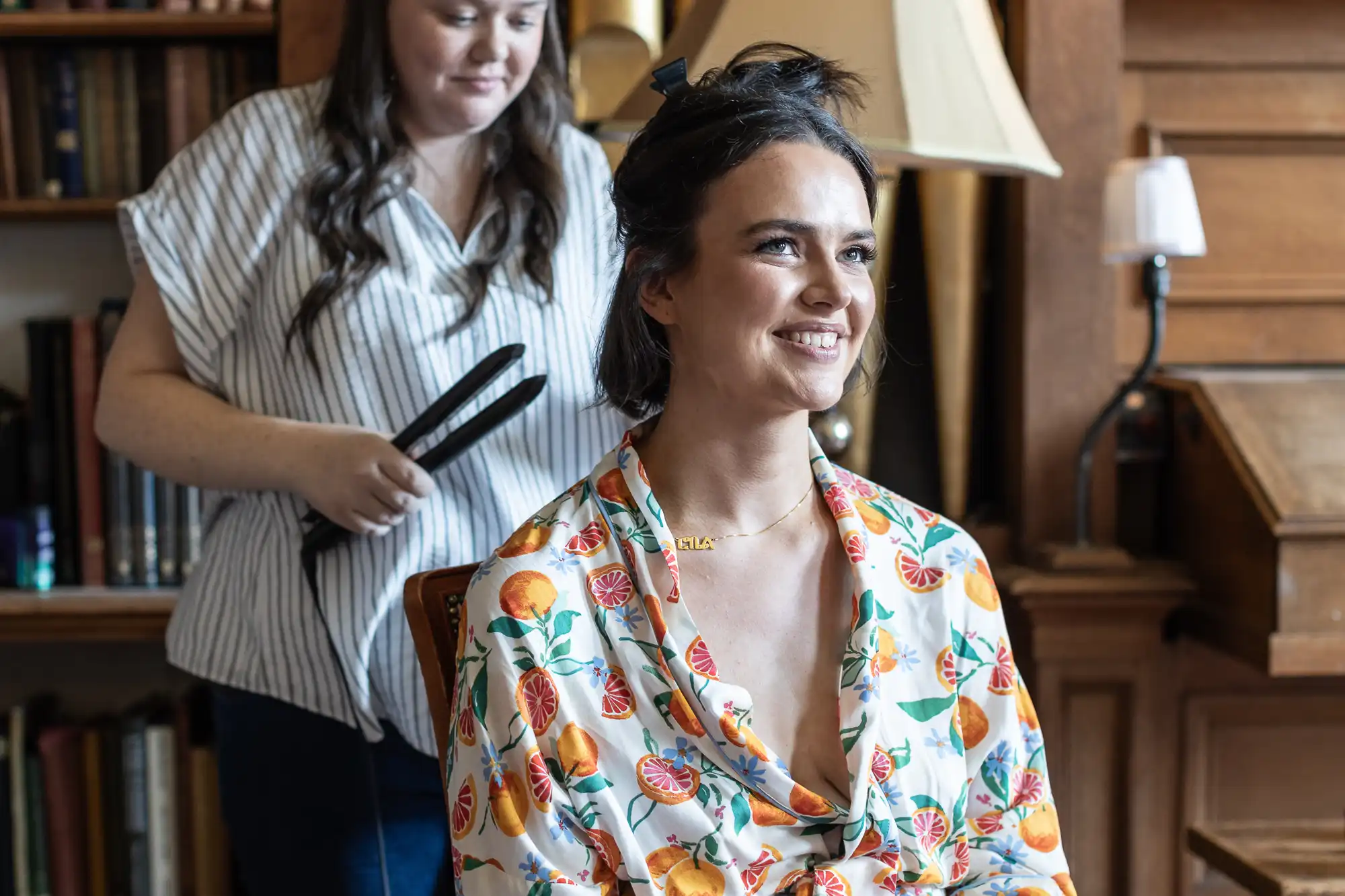 A woman in a fruit-patterned robe smiles while sitting, as another woman styles her hair with a flat iron in a room with bookshelves and a lamp.