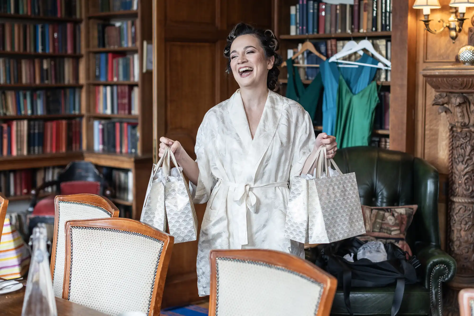 A woman in a robe smiles, holding two patterned gift bags. She stands in a room with wooden bookshelves, colorful dresses hanging, and a dining table with chairs.