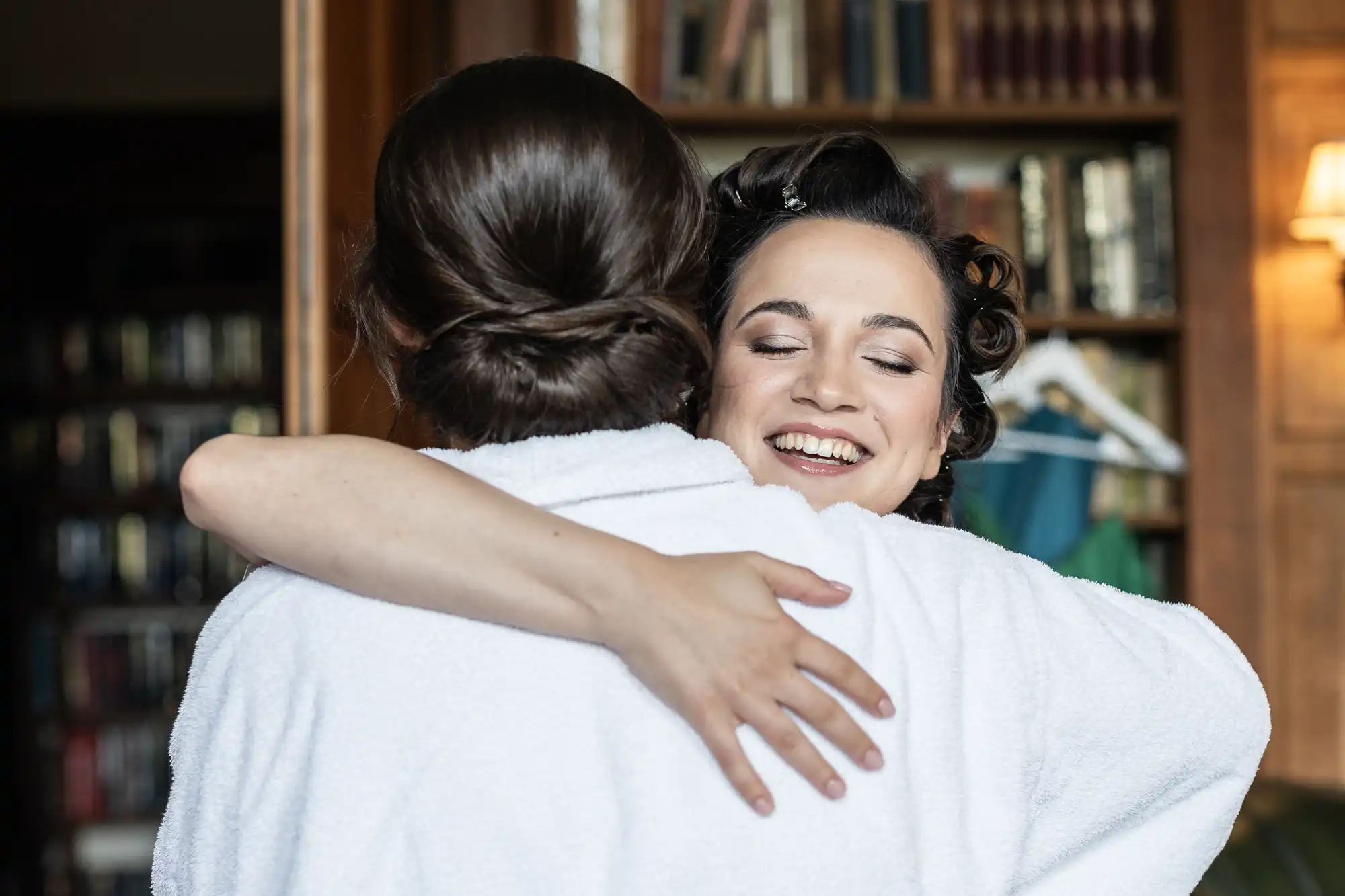 Two people in white robes embrace each other, with one facing away and the other smiling. They are indoors, in a room with bookshelves and warm lighting.