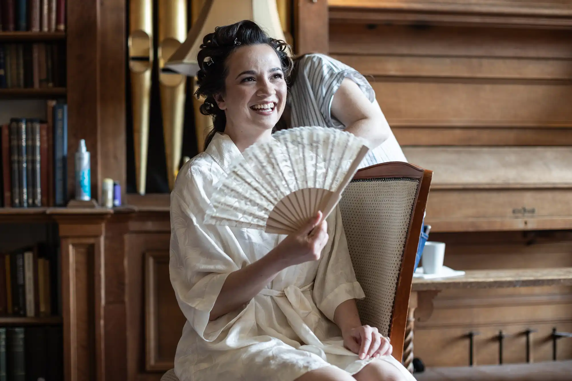 Person in a white robe sitting on a chair, smiling while holding a folding fan. Another person stands nearby, partially visible. Wooden shelves with books are in the background.